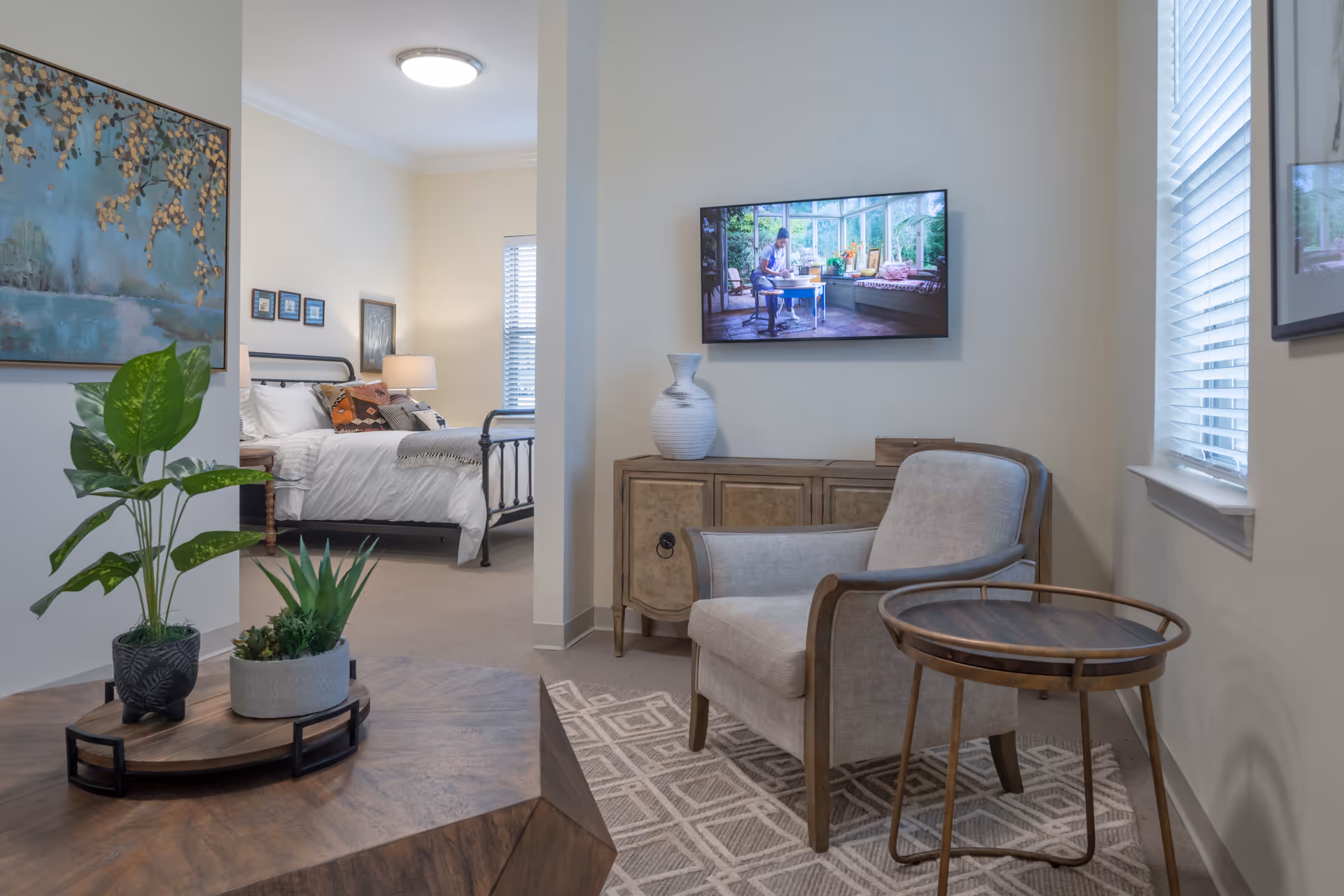 A cozy living area with a beige armchair, a round wooden side table, and a wooden cabinet with a white vase on top. A flat-screen TV is mounted on the wall above the cabinet. To the left, there is a view into a bedroom with a bed, pillows, a bedside table with a lamp, and wall art. The room has neutral tones and natural light coming through the windows with blinds.