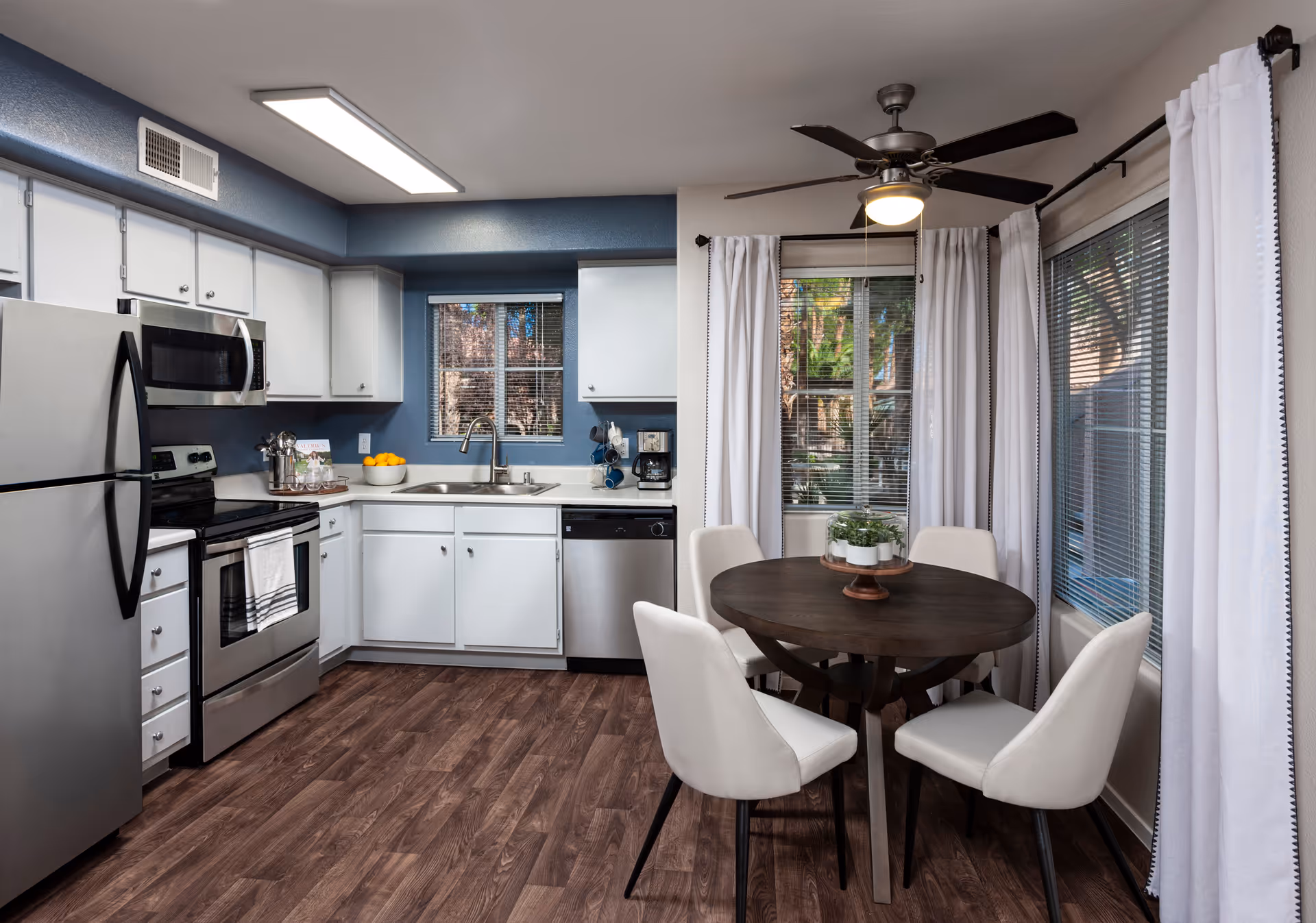 A modern kitchen and dining area with white cabinets, stainless steel appliances including a refrigerator, microwave, stove, and dishwasher. The kitchen has a window above the sink with blinds. Adjacent to the kitchen is a round dark wooden dining table with four white upholstered chairs. The dining area has large windows with white curtains and a ceiling fan with a light fixture.