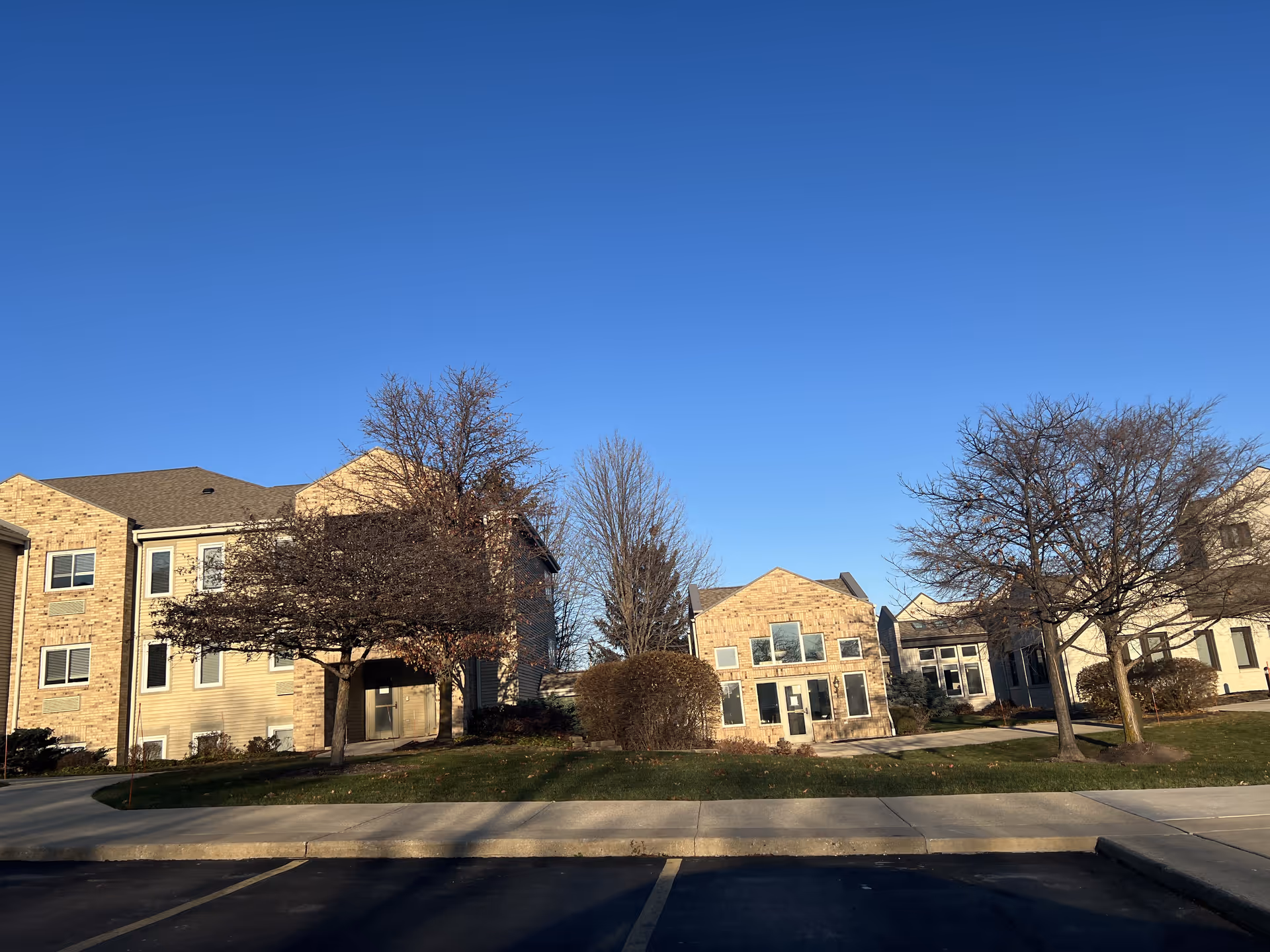 Exterior view of a senior living facility with multiple connected buildings made of brick and siding, surrounded by leafless trees and bushes under a clear blue sky. There is a paved parking area and a sidewalk in the foreground.