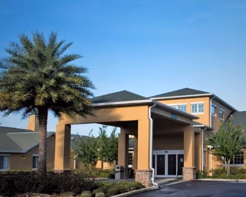 Exterior view of a senior living facility building with a covered entrance supported by columns, surrounded by landscaping including a palm tree and bushes under a clear blue sky.