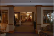 Interior view of a senior living facility showing a hallway leading to a common area with chairs and tables. The space is warmly lit with wooden trim and columns, and there are comfortable upholstered chairs and windows on the sides.