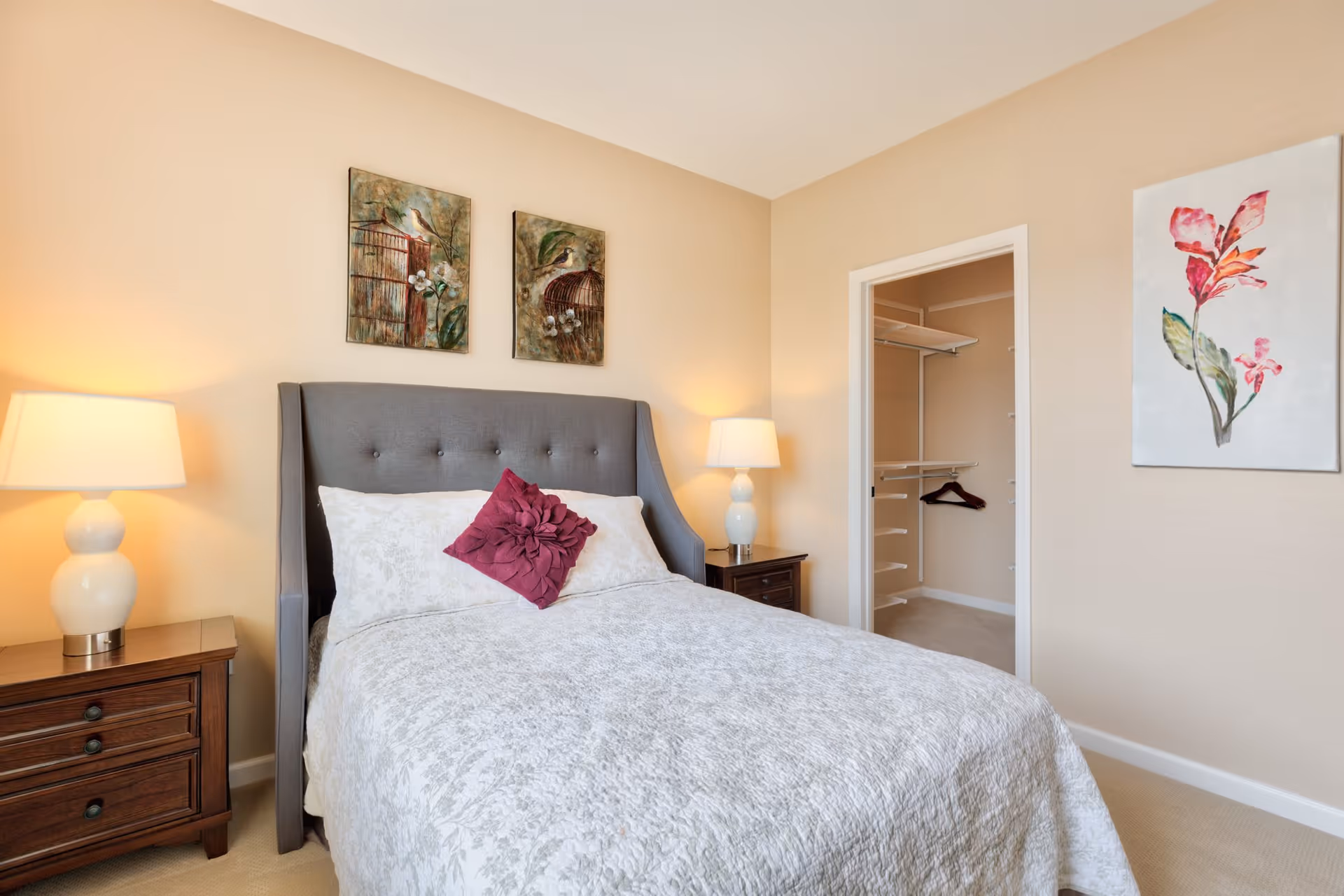 A bedroom with a gray upholstered bed featuring a white patterned bedspread and a decorative purple pillow. There are two wooden nightstands with white lamps on either side of the bed. Above the bed are two framed artworks depicting birds and birdcages. To the right, there is an open doorway leading to a walk-in closet with shelving and a single hanger. A floral painting is hung on the wall next to the closet.