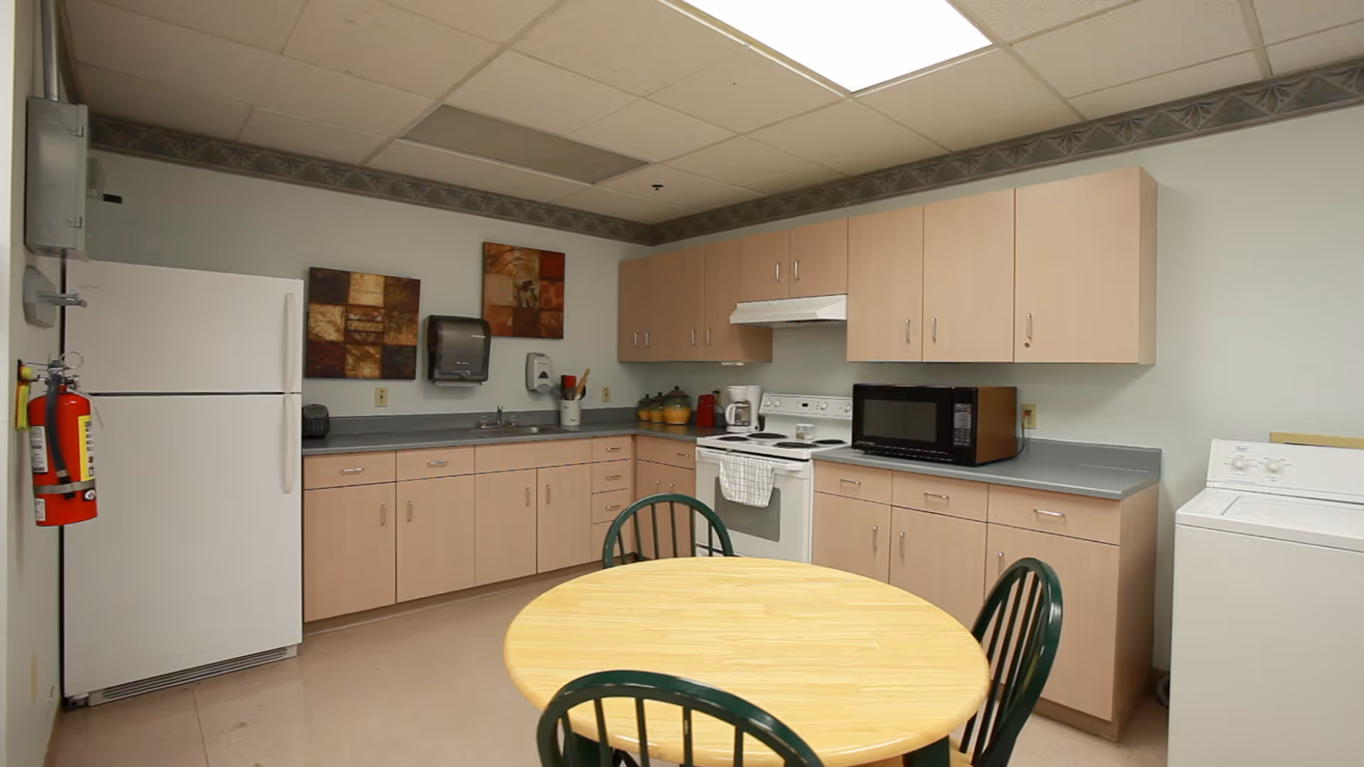A clean kitchen area in a senior living facility with light wood cabinets, a white refrigerator, stove, microwave, and washing machine. There is a round wooden table with four green chairs in the center. The walls are light-colored with two decorative square wall hangings above the sink area.