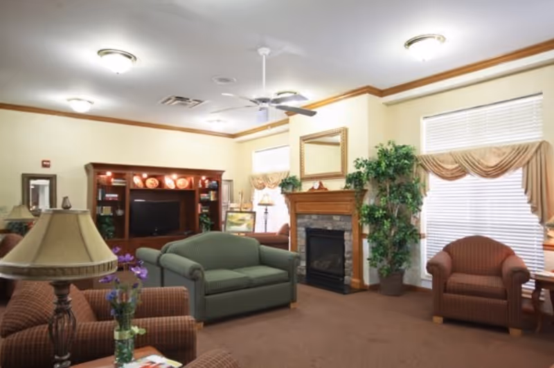 A cozy living room in a senior living facility with a green loveseat, two brown armchairs, a wooden entertainment center with a TV, a fireplace with a stone surround and wooden mantle, potted plants, and windows with beige valances and blinds.