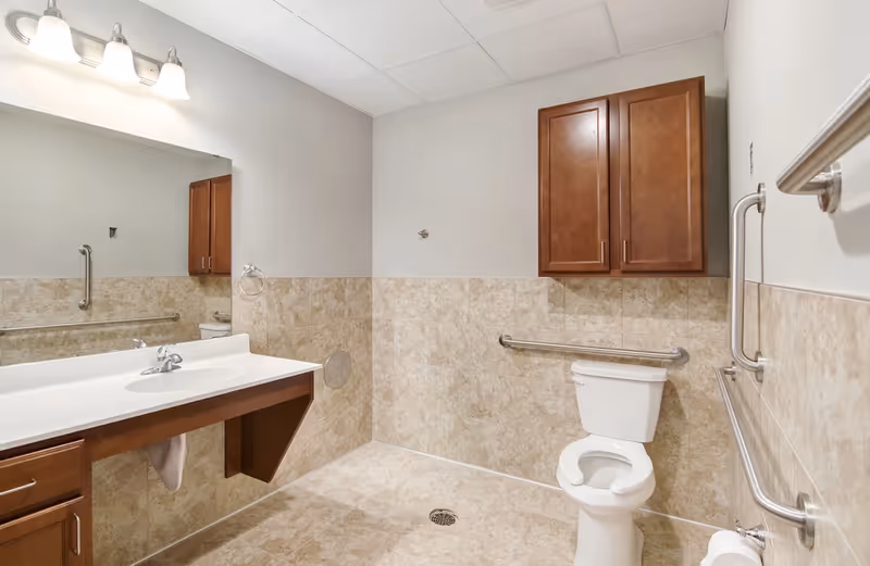 A spacious bathroom with beige tiled walls and floor, featuring a white toilet with grab bars on both sides, a large mirror above a white sink with a wooden cabinet underneath, and a wooden wall cabinet above the toilet. The bathroom has a ceiling light fixture with three bulbs.