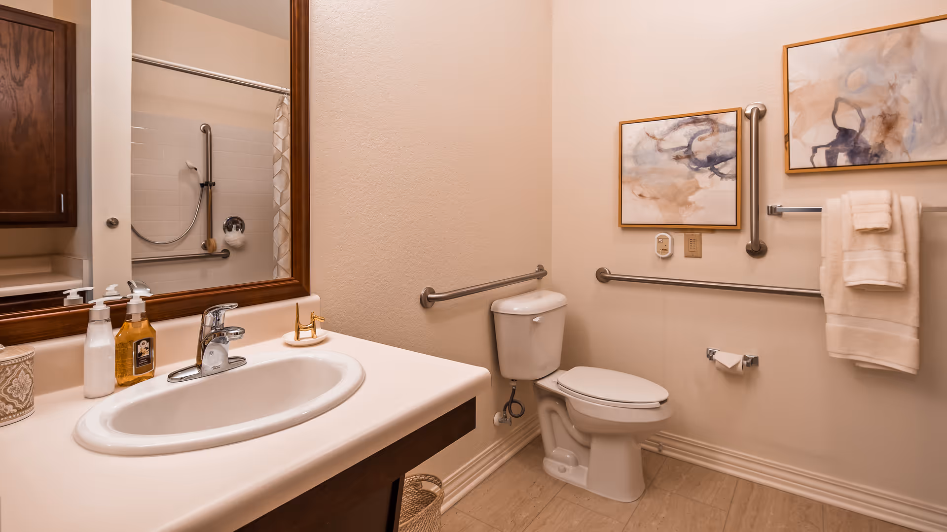 A clean and accessible bathroom with a white sink and countertop, a large mirror above the sink, a toilet with grab bars on the walls, a shower with a curtain and grab bars, two framed abstract paintings on the wall, and neatly folded towels hanging on a towel rack.