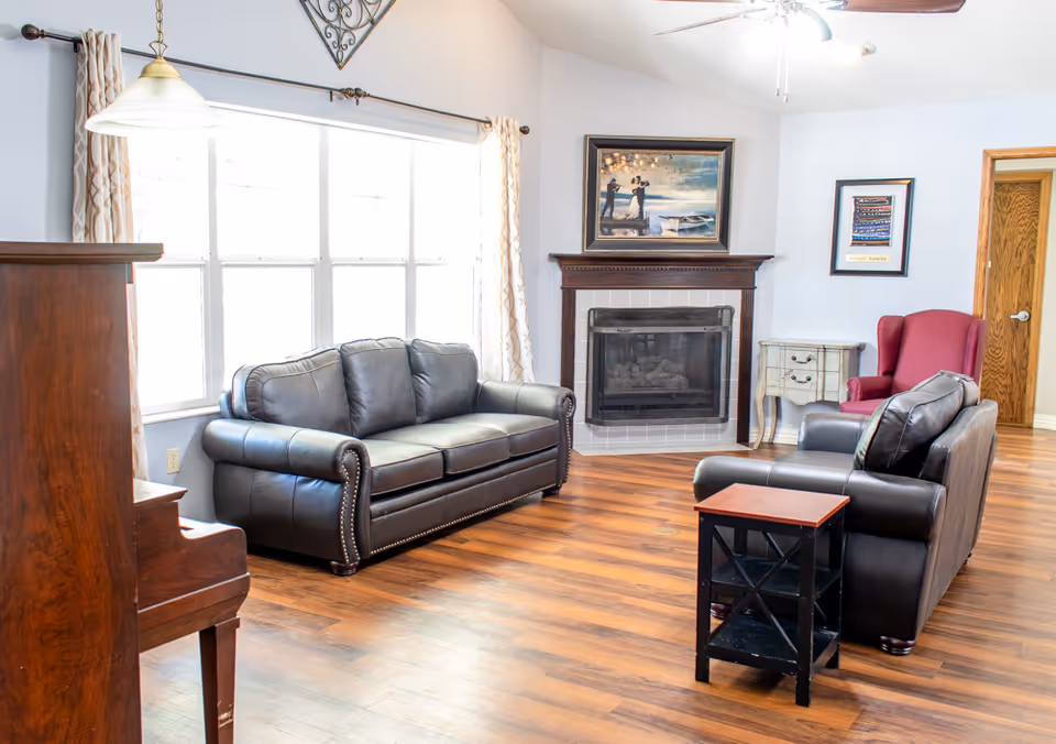 A bright living room with wooden flooring featuring two black leather sofas, a red armchair, a small black side table, a fireplace with a framed painting above it, a wooden piano on the left, and large windows with patterned curtains letting in natural light.