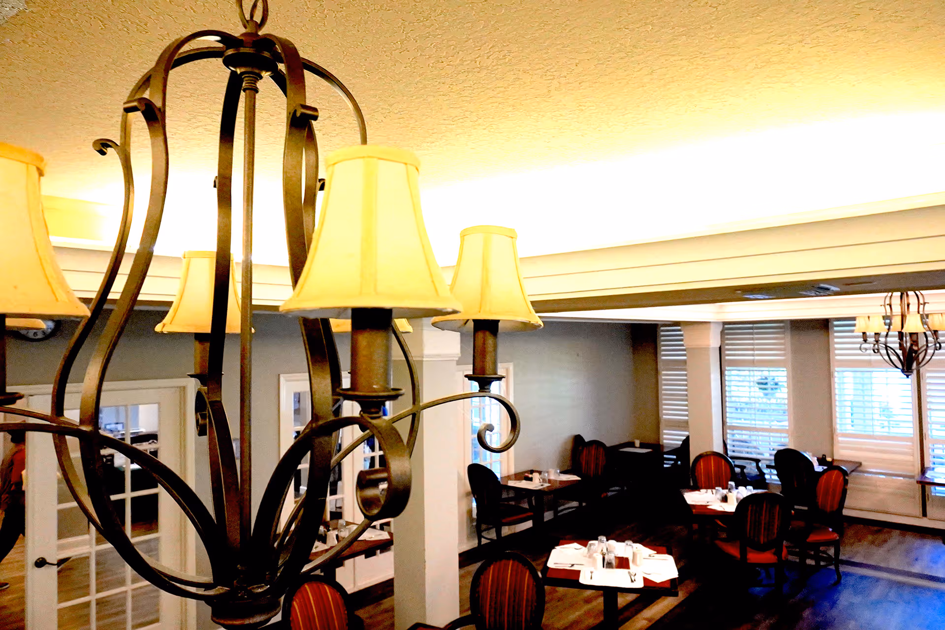Interior view of a dining room with multiple tables set with white napkins, glasses, and silverware. The room features wooden floors, large windows with blinds, and decorative chandeliers with lampshades hanging from the ceiling.