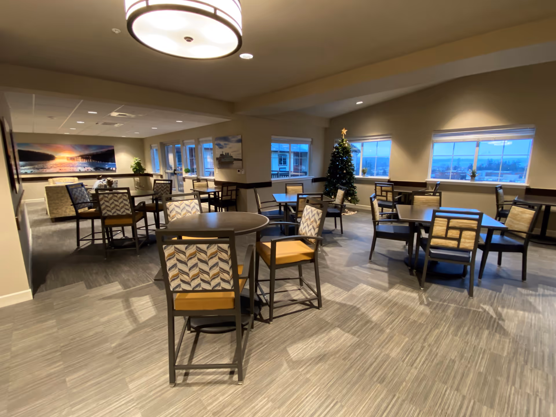 A spacious dining area with multiple tables and chairs arranged neatly. The room has large windows letting in natural light, a decorated Christmas tree in the corner, and framed landscape artwork on the walls. The flooring is a mix of carpet and tile, and a large ceiling light fixture is visible.