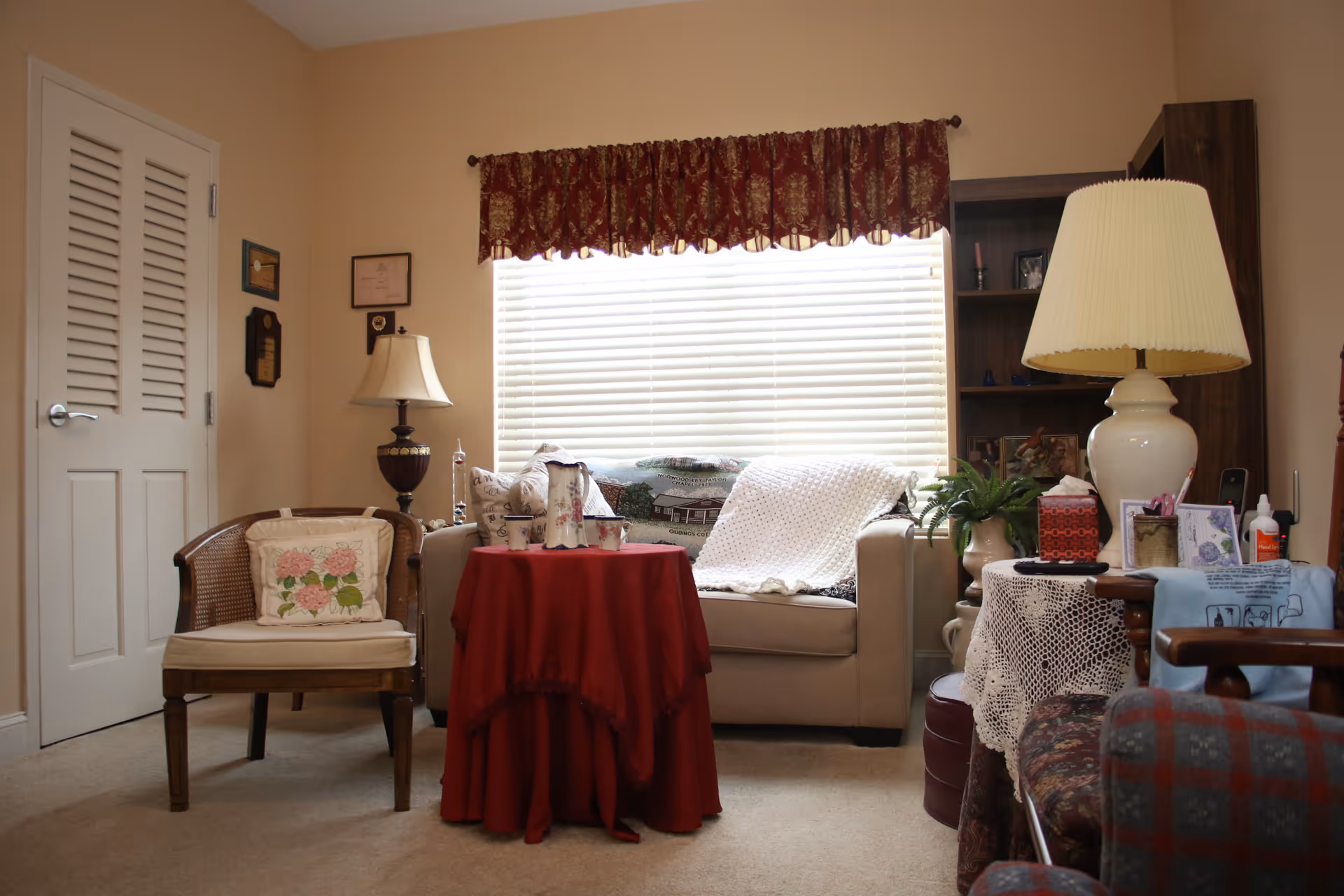 Cozy living room with a sofa, armchairs, lamps, and a small round table draped in a red cloth in front of a window with blinds.