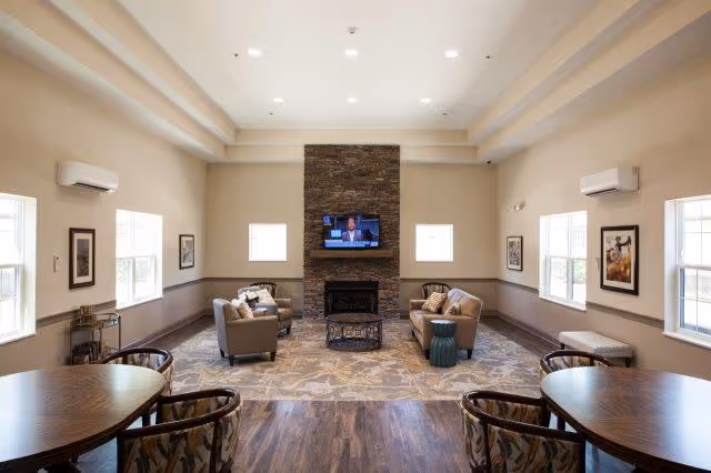 A spacious living room with a high ceiling and recessed lighting. The room features a stone fireplace with a flat-screen TV mounted above it. There are two beige sofas and two armchairs arranged around the fireplace, with a small green side table. The room has several windows letting in natural light, framed artwork on the walls, and two round wooden tables with patterned chairs in the foreground. The floor is a combination of wood and carpet.
