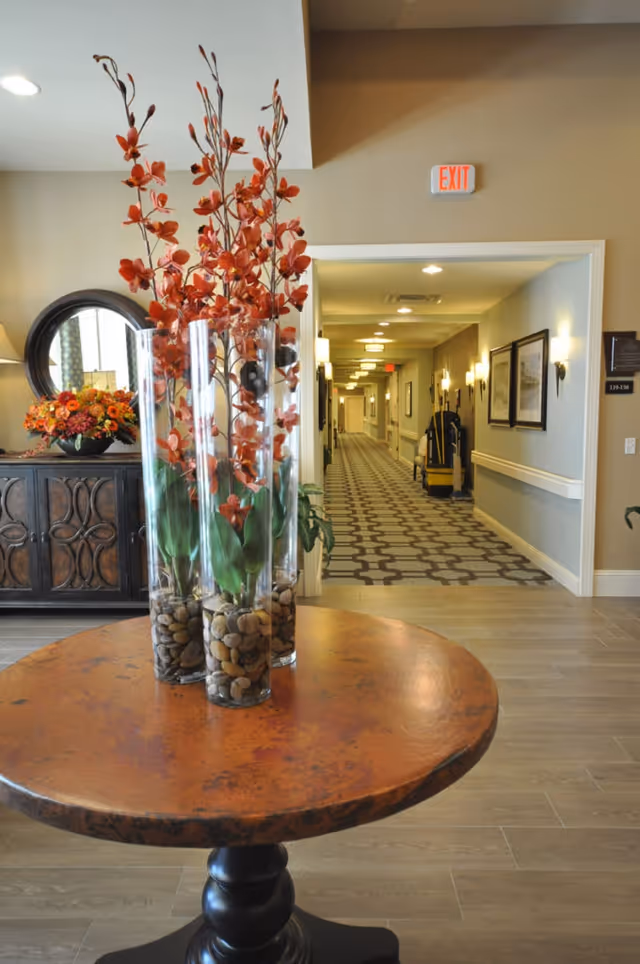 Round entry table with tall glass vases of orange flowers in a lobby opening onto a long hallway with an exit sign.