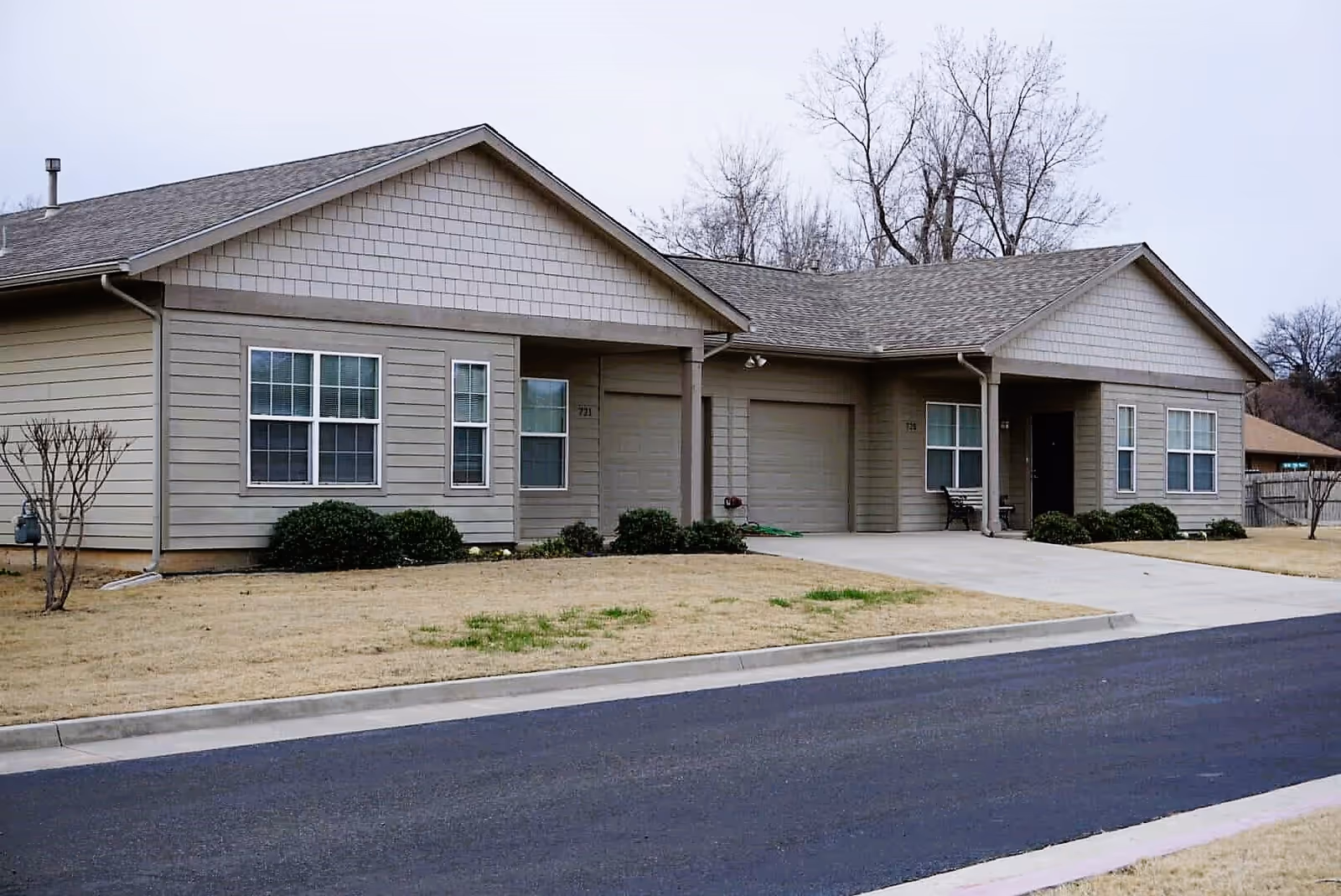 Exterior view of a single-story residential building with beige siding and a gray shingled roof. The building has multiple windows, two garage doors, and a driveway. There are small bushes and a leafless tree in the front yard with a paved road in the foreground. The sky is overcast.