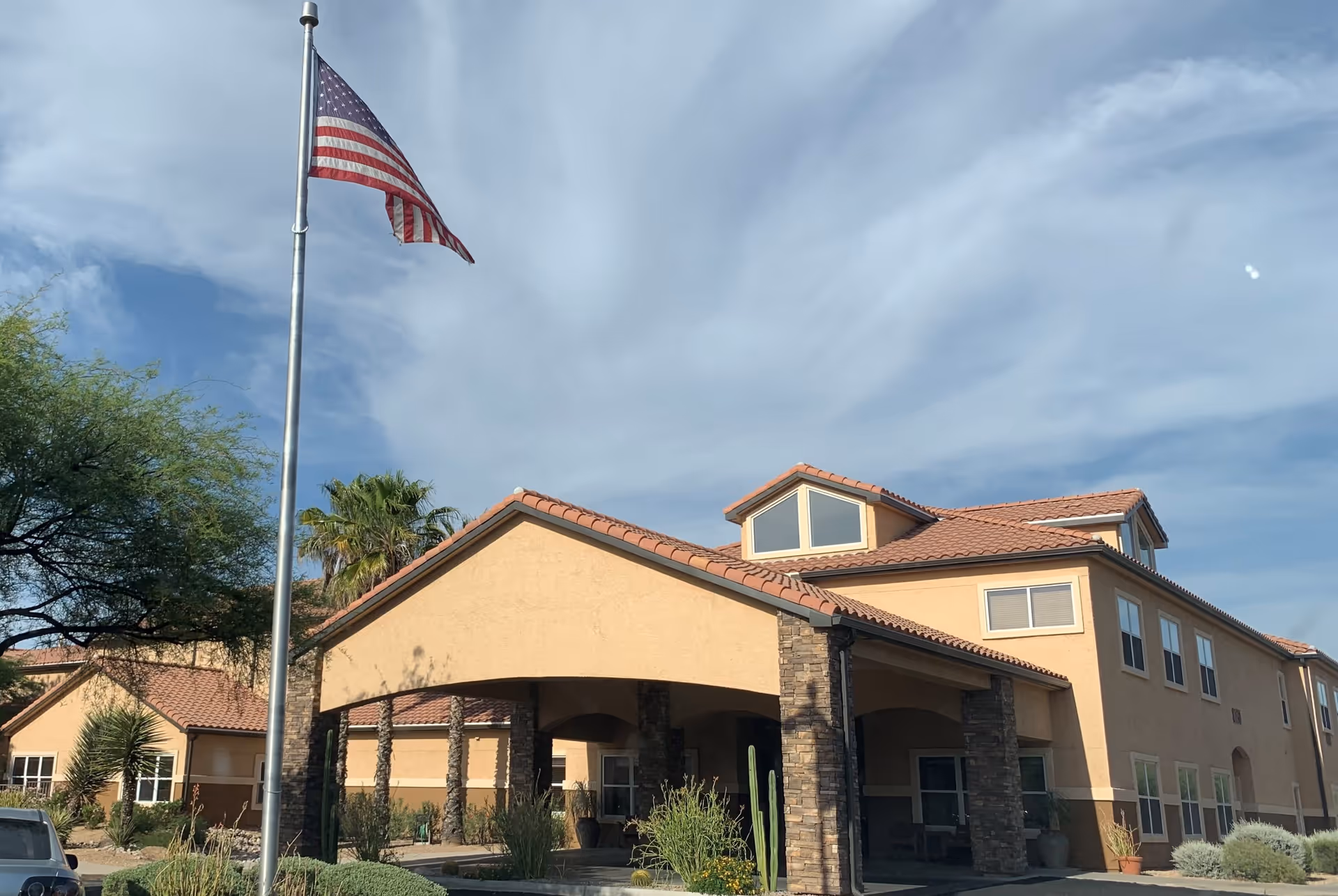 Exterior view of Sierra Del Sol Assisted Living facility with a beige stucco building featuring a tiled roof, multiple windows, and a covered entrance supported by stone pillars. An American flag on a tall flagpole is prominently displayed in front of the building, with desert landscaping including cacti and shrubs visible around the entrance.