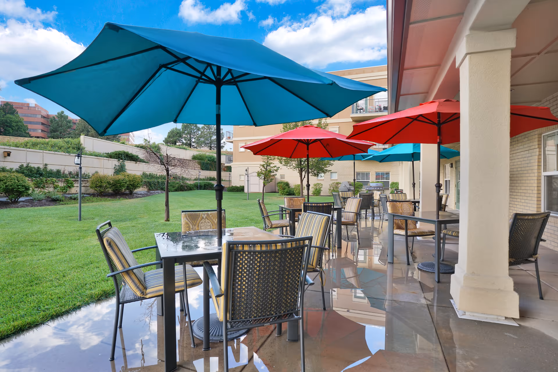 Outdoor patio area with several tables and chairs under large blue and red umbrellas. The patio is adjacent to a building with columns and overlooks a grassy area with bushes and trees. The sky is partly cloudy.