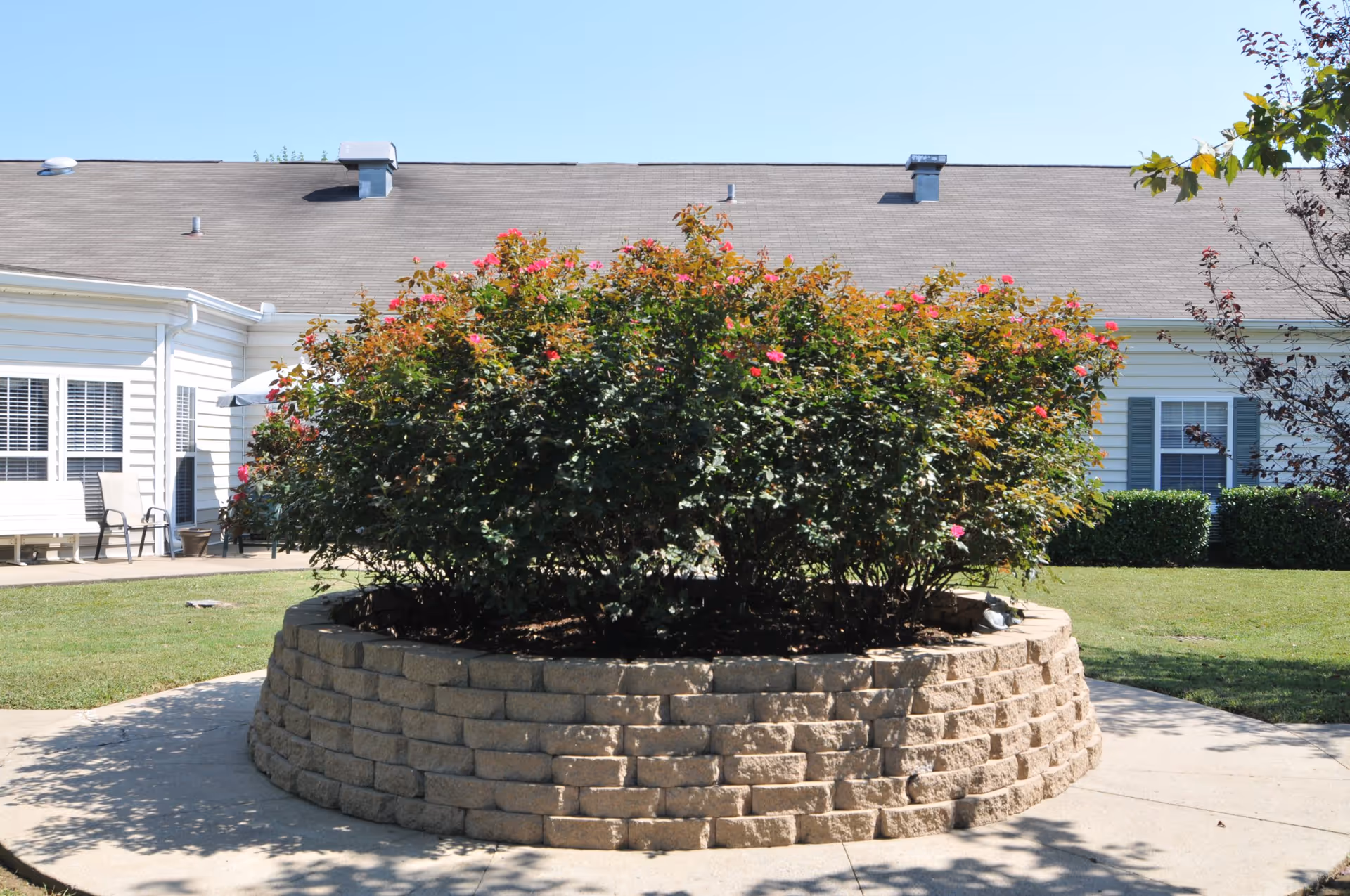 A circular raised flower bed made of beige stone bricks filled with green bushes and pink flowers, situated on a concrete patio area. In the background, there is a white building with windows and a gray shingled roof under a clear blue sky.