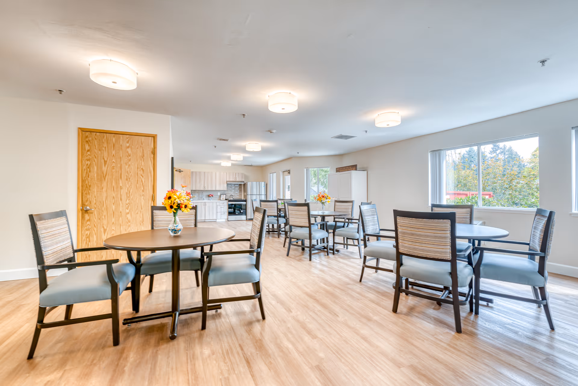 A bright and spacious dining area in a senior living facility with round tables and chairs arranged neatly. Each table has a small vase with colorful flowers. The room has large windows letting in natural light, light wood flooring, and modern ceiling lights. In the background, there is a kitchen area with cabinets, a refrigerator, and a stove.