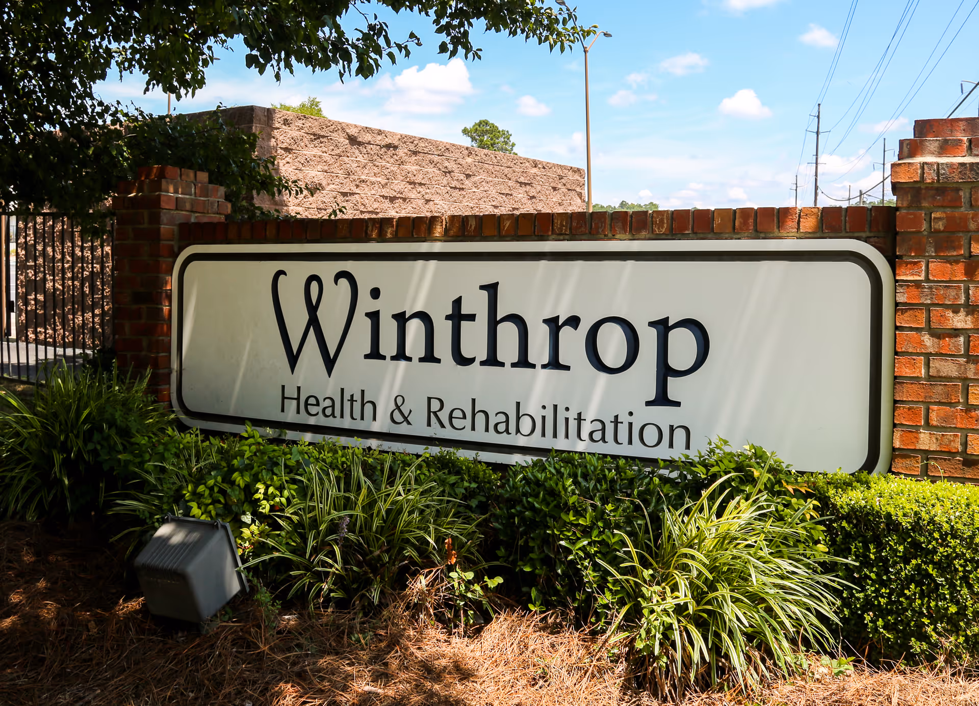 Outdoor sign for Winthrop Health & Rehabilitation mounted on a brick wall with greenery and plants in front, under a partly cloudy blue sky.