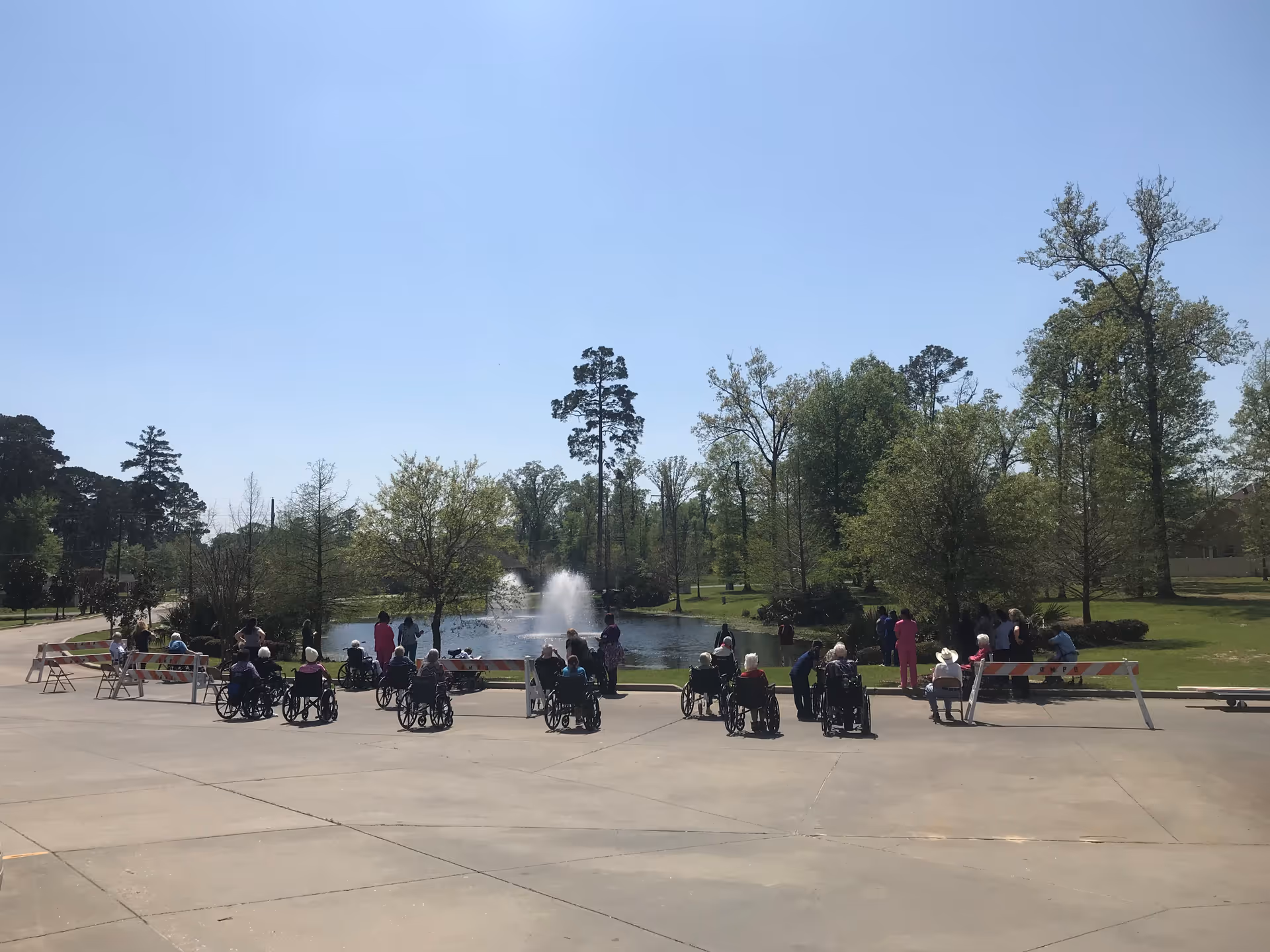 A group of elderly people, many in wheelchairs, gathered outside near a pond with a water fountain. They are accompanied by caregivers and are enjoying a sunny day surrounded by trees and greenery.