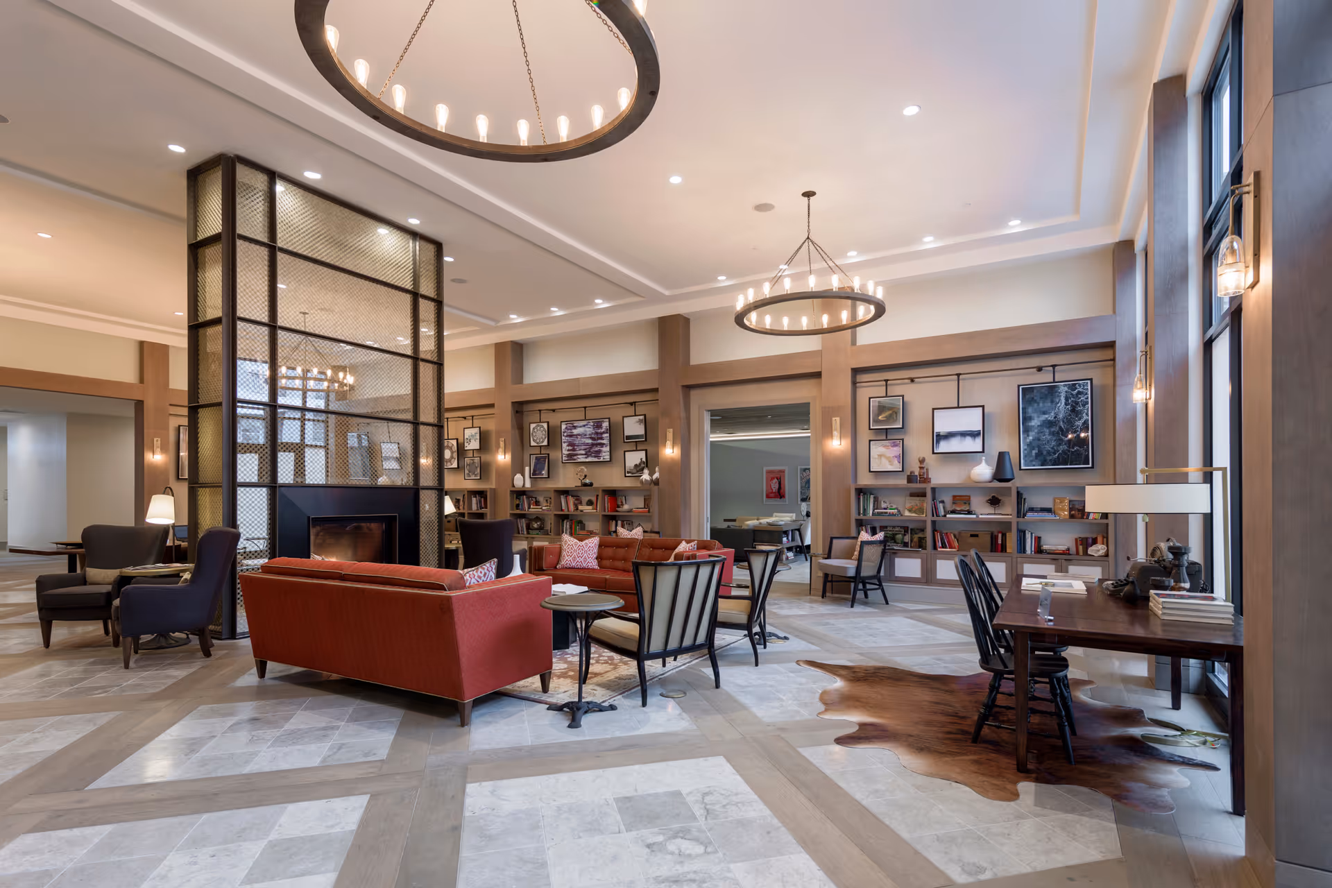A spacious and well-lit living room area with modern decor featuring a central fireplace surrounded by a metal mesh screen. The room has multiple seating options including red sofas, armchairs, and a wooden desk with chairs near large windows. The walls are adorned with framed artwork and built-in bookshelves filled with books and decorative items. Two large circular chandeliers hang from the ceiling, illuminating the space.
