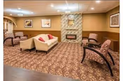 A cozy living room area with patterned carpet, a beige sofa with red cushions, several patterned armchairs, a stone fireplace with a clock above it, and framed artwork on the walls.