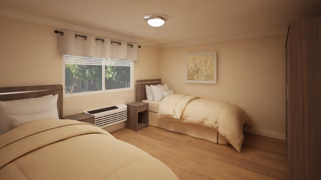 A cozy bedroom with two single beds featuring beige bedding and white pillows. Between the beds is a small wooden nightstand. A window with white curtains lets in natural light, showing green foliage outside. The room has wooden flooring, a ceiling light, a framed artwork on the wall, and a wooden wardrobe on the right side.