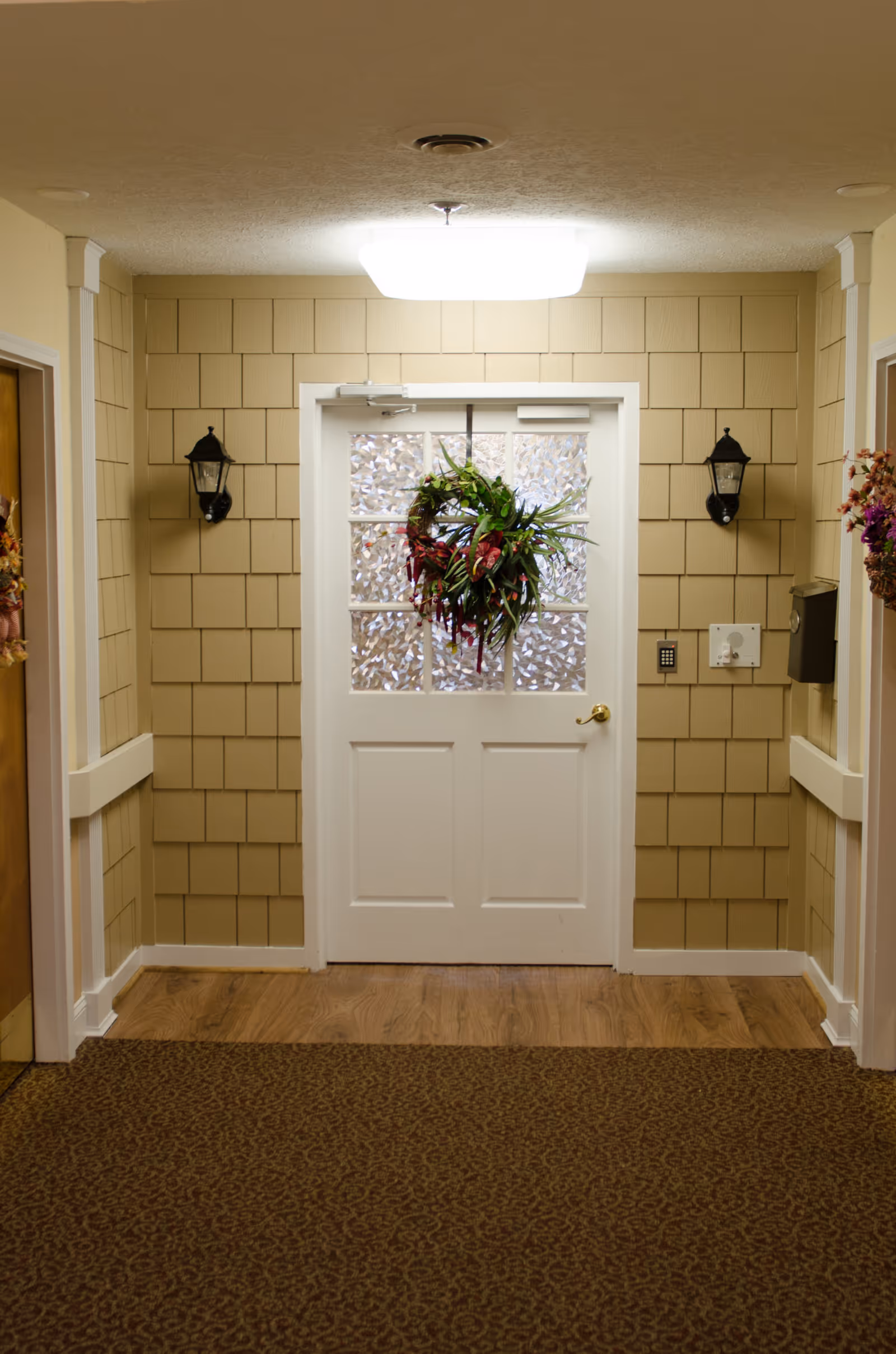 Interior hallway with beige shingle-style walls and a white door with frosted glass panels decorated with a green and red wreath. Two black wall lanterns flank the door, and there are floral decorations on the walls near two wooden doors on either side. The floor is carpeted with a patterned brown carpet, and there is a wooden floor section in front of the door.
