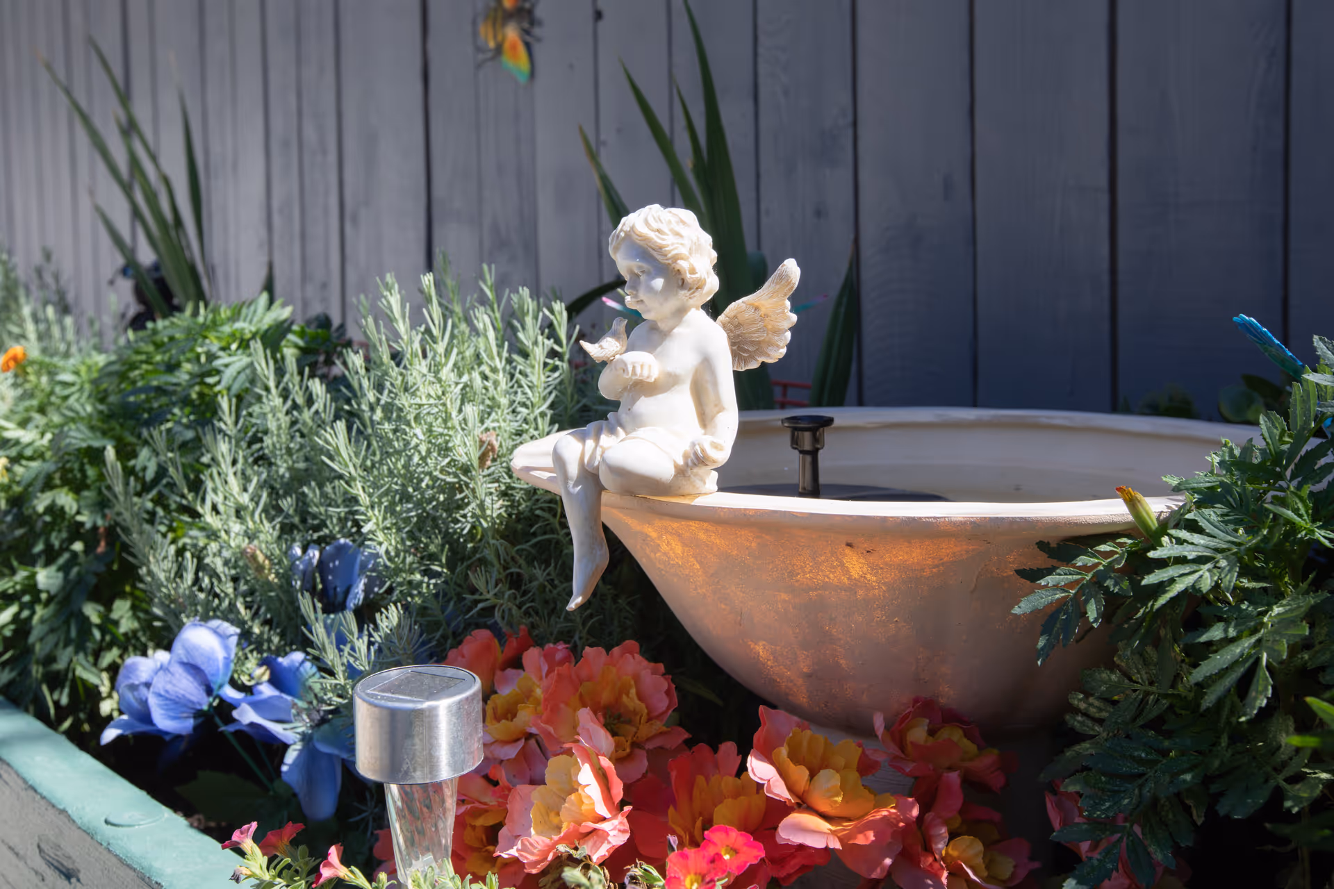 A garden scene featuring a small white cherub statue with wings sitting on the edge of a birdbath surrounded by colorful flowers and green plants, with a wooden fence in the background.
