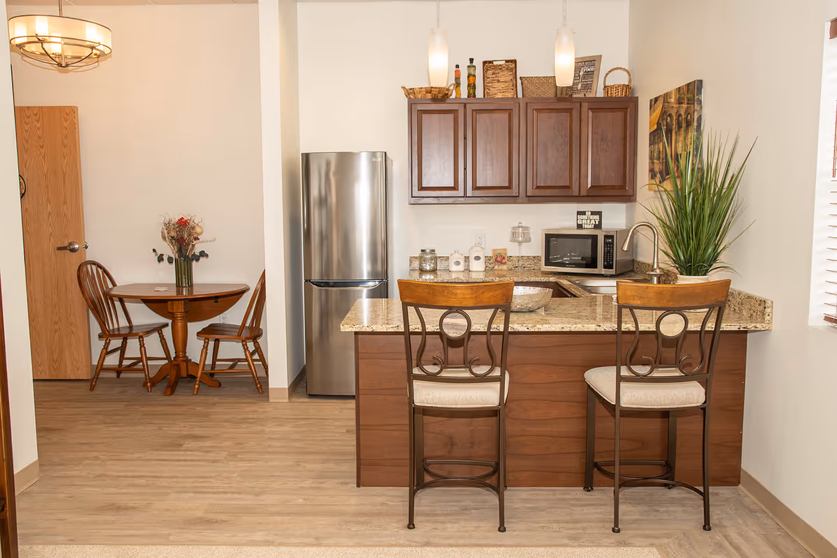 A cozy kitchen and dining area featuring a granite countertop with two high chairs, wooden cabinets, a stainless steel refrigerator, a microwave, and a sink. In the background, there is a small round wooden dining table with two chairs and a vase with flowers. The room has light-colored walls and wood flooring.