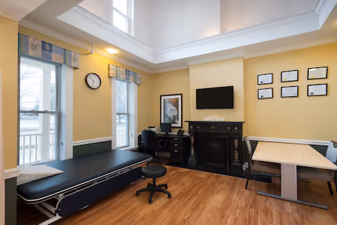 A well-lit medical examination room with a black examination table on the left, a black swivel stool in front, a desk with a chair and computer monitor near a fireplace, and a table with bench seating on the right. The walls are painted yellow with white trim, and several framed certificates and a clock are displayed on the walls.