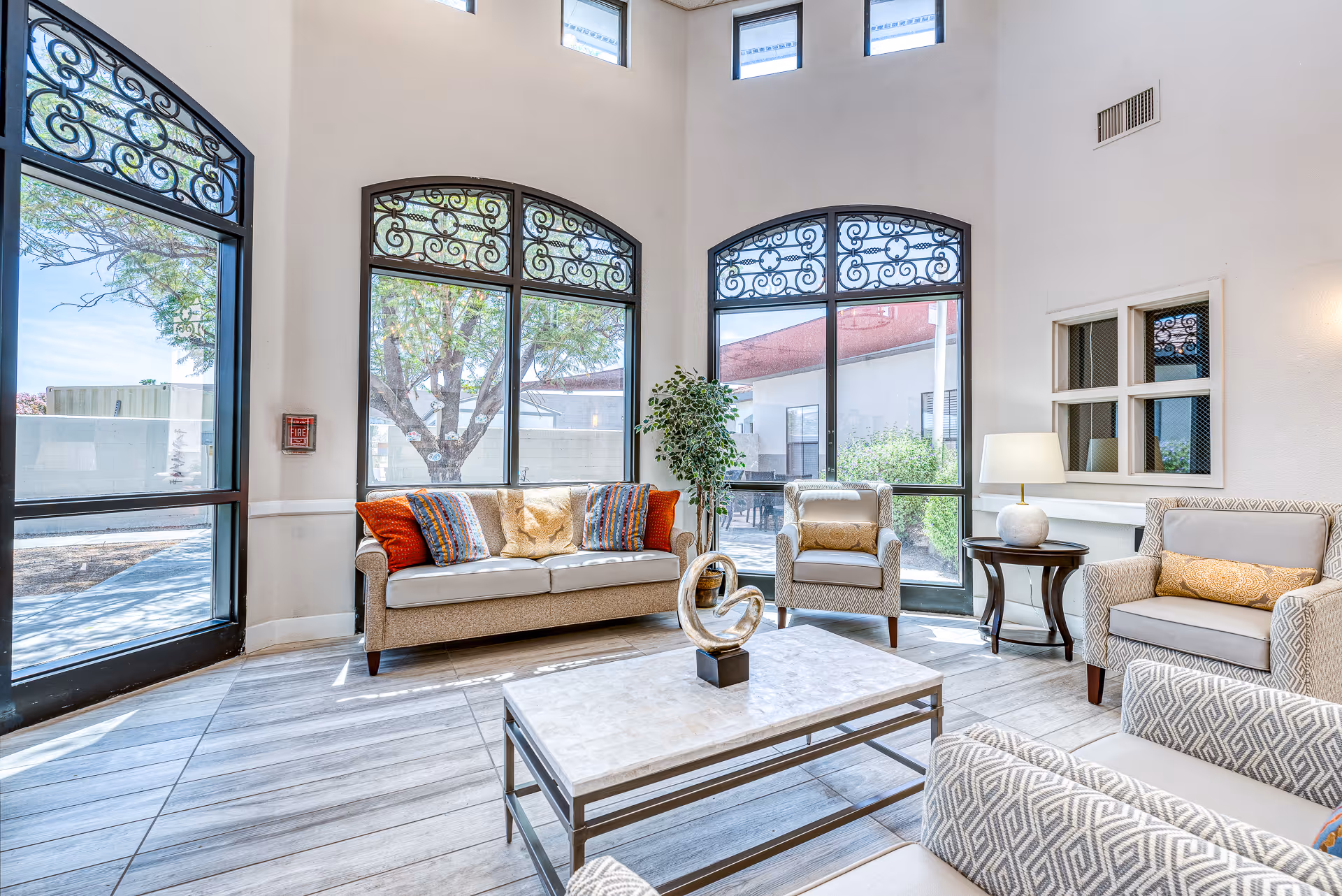Bright and airy living room with large arched windows featuring decorative wrought iron designs. The room contains a beige sofa with colorful cushions, two patterned armchairs with yellow pillows, a marble-top coffee table with a modern sculpture, a side table with a lamp, and a potted plant near the windows. The floor is tiled with light wood-like tiles.