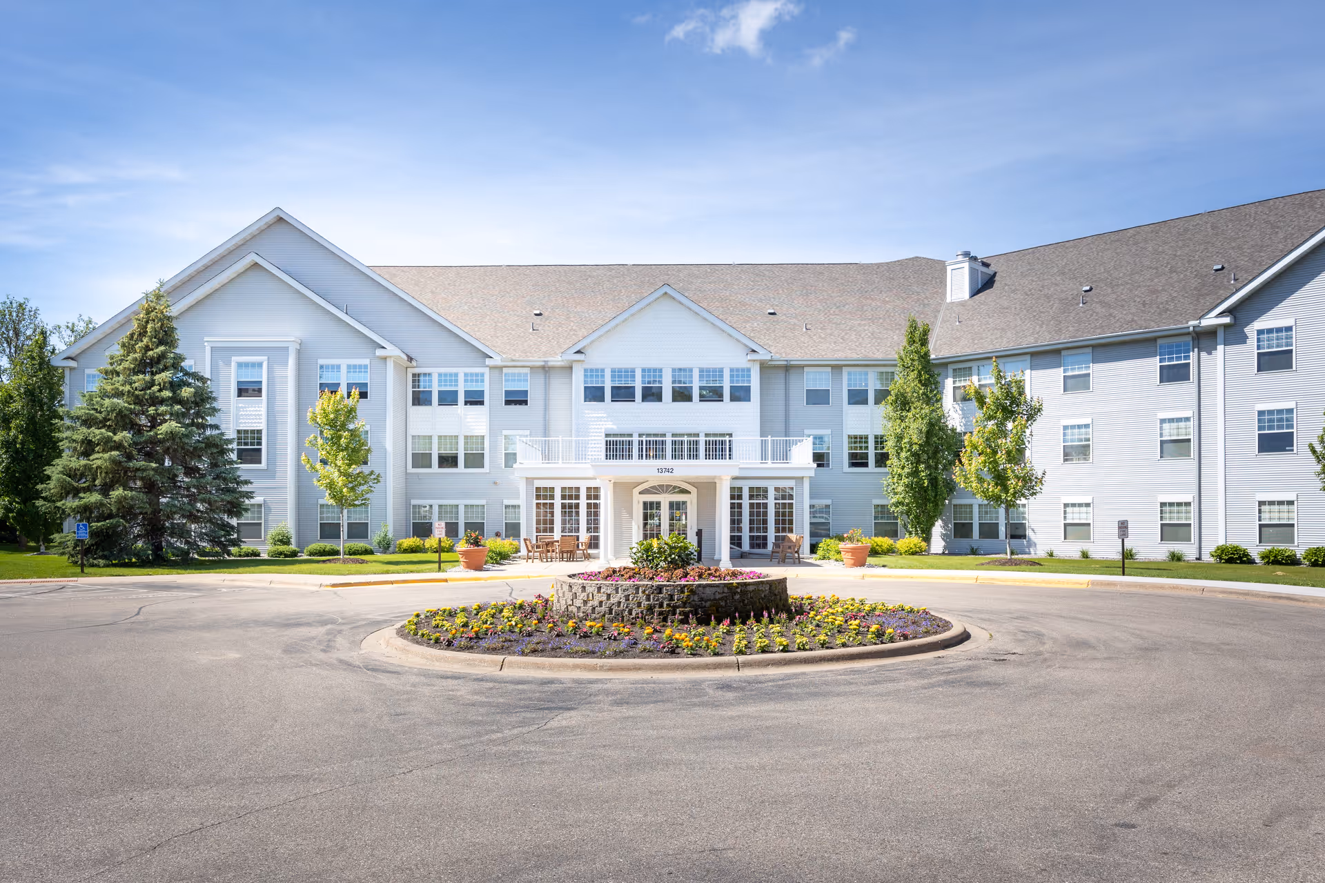 Front exterior view of a large, three-story senior living facility named The Farmstead, featuring a circular driveway with a landscaped flower bed in the center, surrounded by trees and greenery under a clear blue sky.