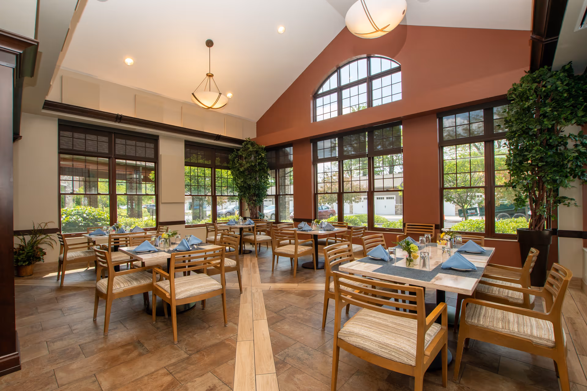 A bright dining room with multiple wooden tables and chairs arranged neatly. Each table is set with blue napkins, glasses, and small flower arrangements. Large windows with dark frames allow natural light to fill the room, and there are tall green plants in the corners. The ceiling is vaulted with two hanging light fixtures.