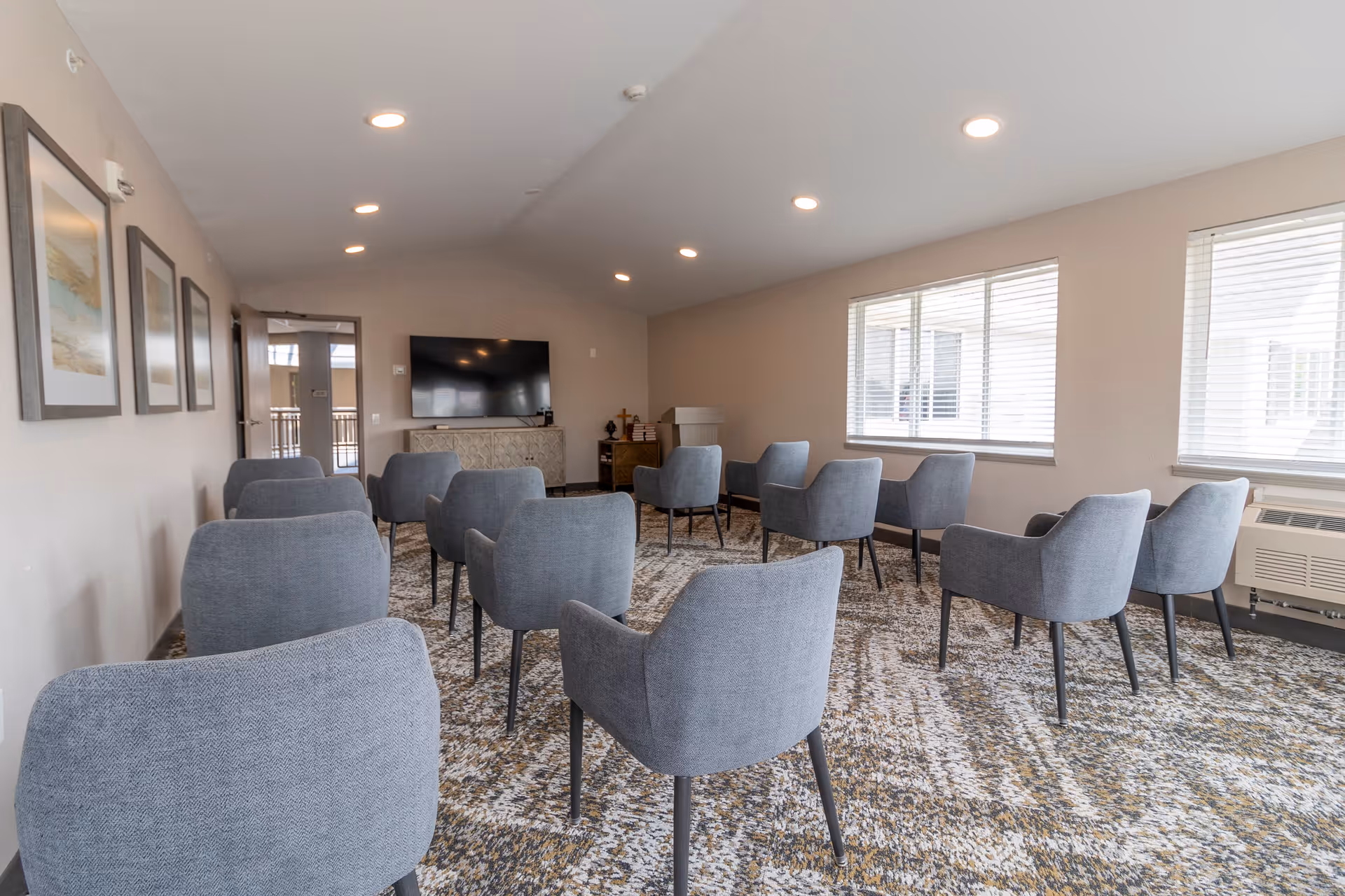 A bright room with multiple gray upholstered chairs arranged in rows facing a large flat-screen TV mounted on the wall. The room has beige walls, three framed pictures on the left wall, large windows with blinds on the right, and a patterned carpeted floor. There is a cabinet under the TV and some decorative items on it.