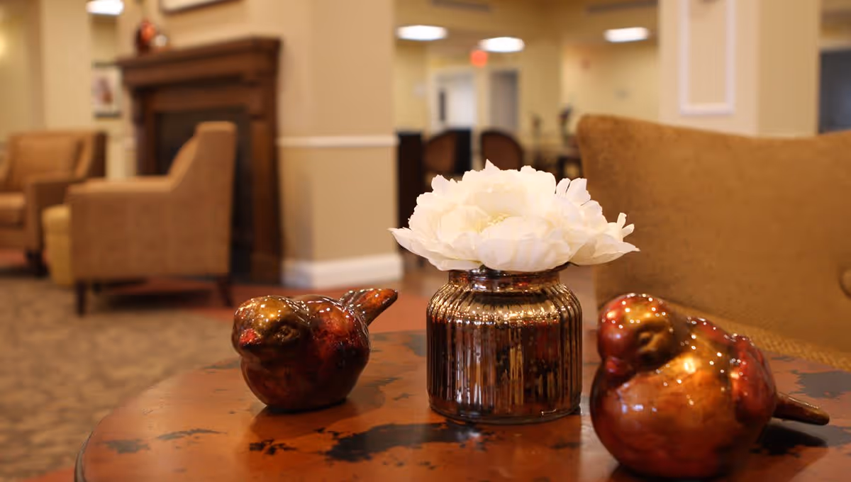 Decorative vase with white flowers and two ceramic bird figurines on a table in a lounge seating area.