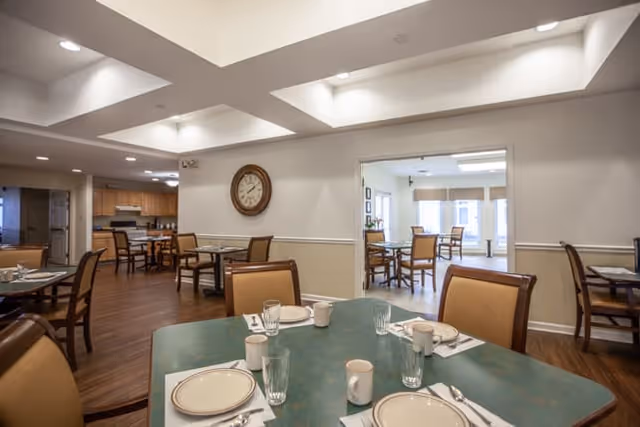 Interior view of a dining room in a senior living facility with multiple tables set with plates, cups, glasses, and silverware. The room has wooden flooring, beige walls with white trim, and a large clock on the wall. There is an adjacent room visible through an open doorway, also furnished with dining tables and chairs.