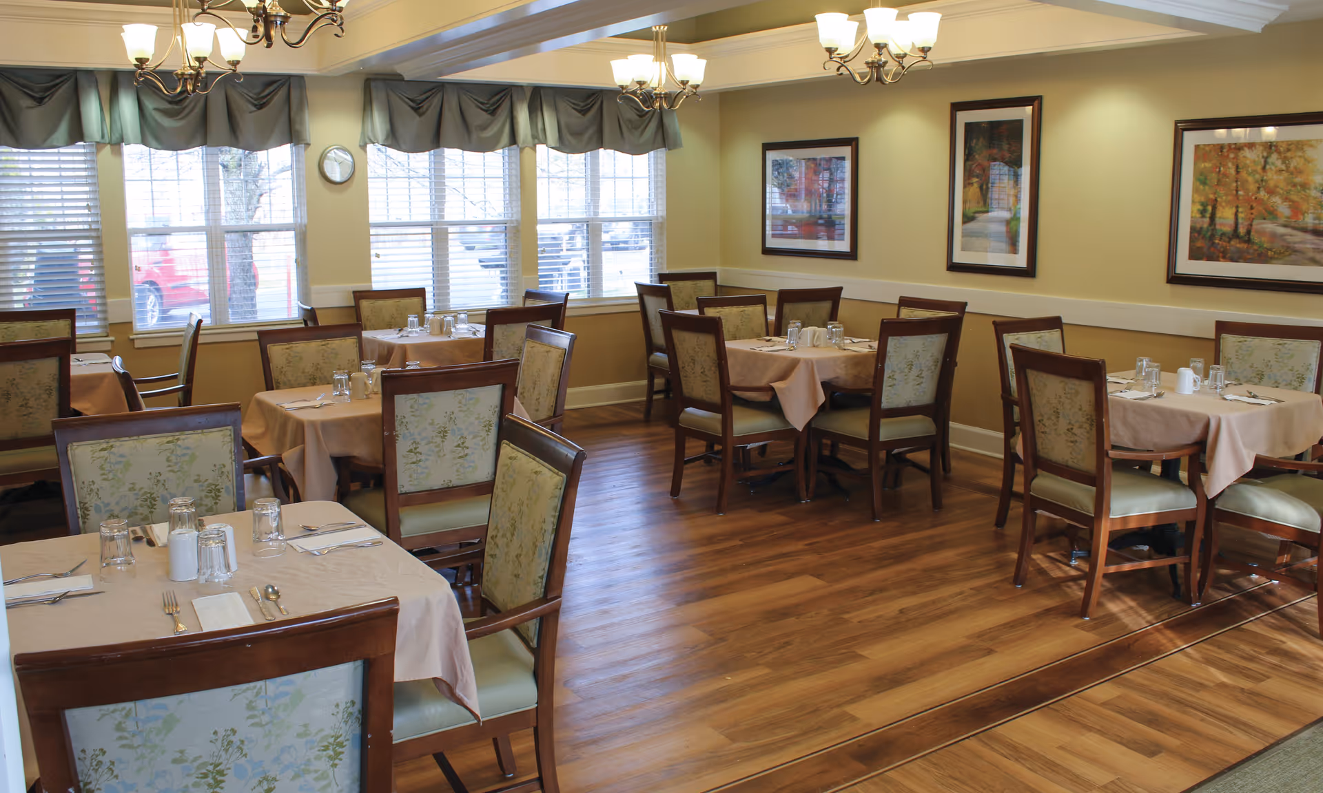 A dining room with several tables covered in beige tablecloths, each set with glasses, napkins, and silverware. The chairs have wooden frames with floral upholstered backs and seats. The room has large windows with blinds and valances, wooden flooring, and framed artwork on the walls. Two chandeliers with multiple lights hang from the ceiling.