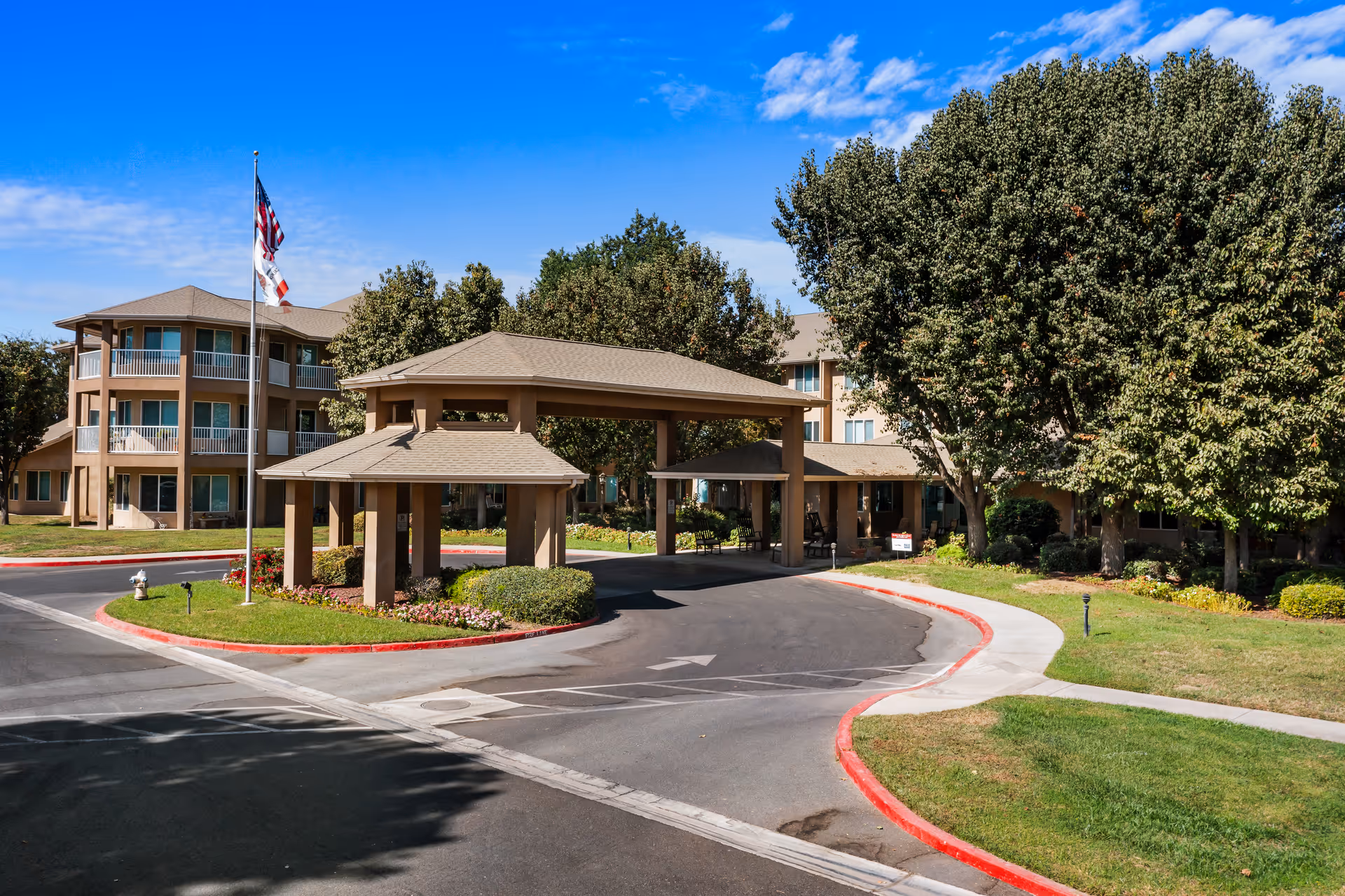Front entrance and porte-cochere of a three-story senior living community with a flagpole, landscaped driveway and trees.