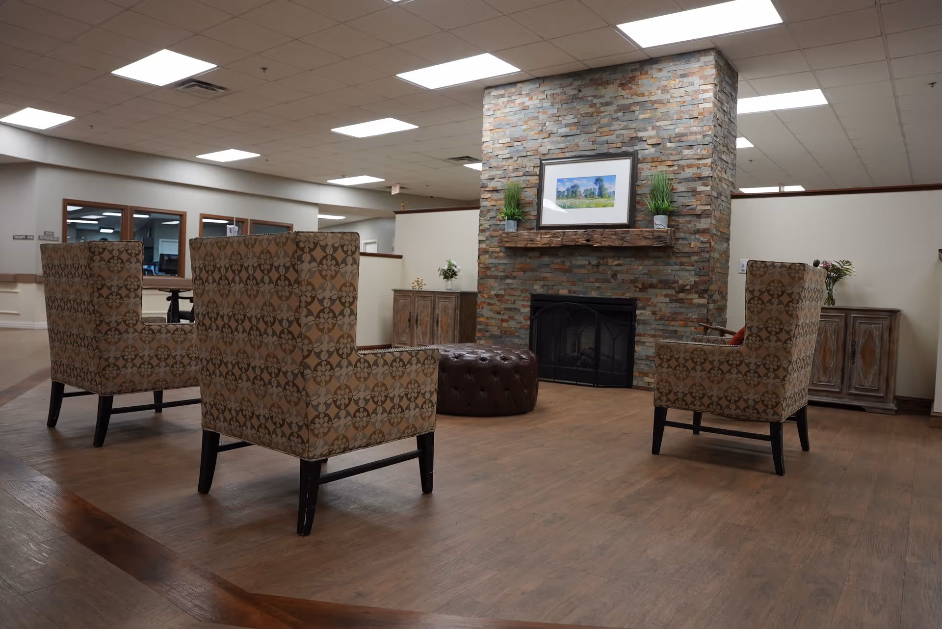 A cozy seating area in a senior living facility with three patterned armchairs arranged around a round, tufted ottoman in front of a stone fireplace. The fireplace has a wooden mantel with two small potted plants and a framed landscape picture above it. The room has wooden flooring, white walls, and a drop ceiling with fluorescent lighting. There are wooden cabinets with decorative items on either side of the fireplace.