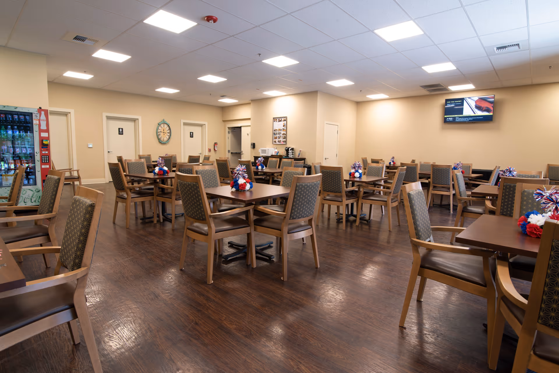 Spacious dining room in a senior living facility with multiple wooden tables and chairs, a vending machine, and a wall-mounted TV.