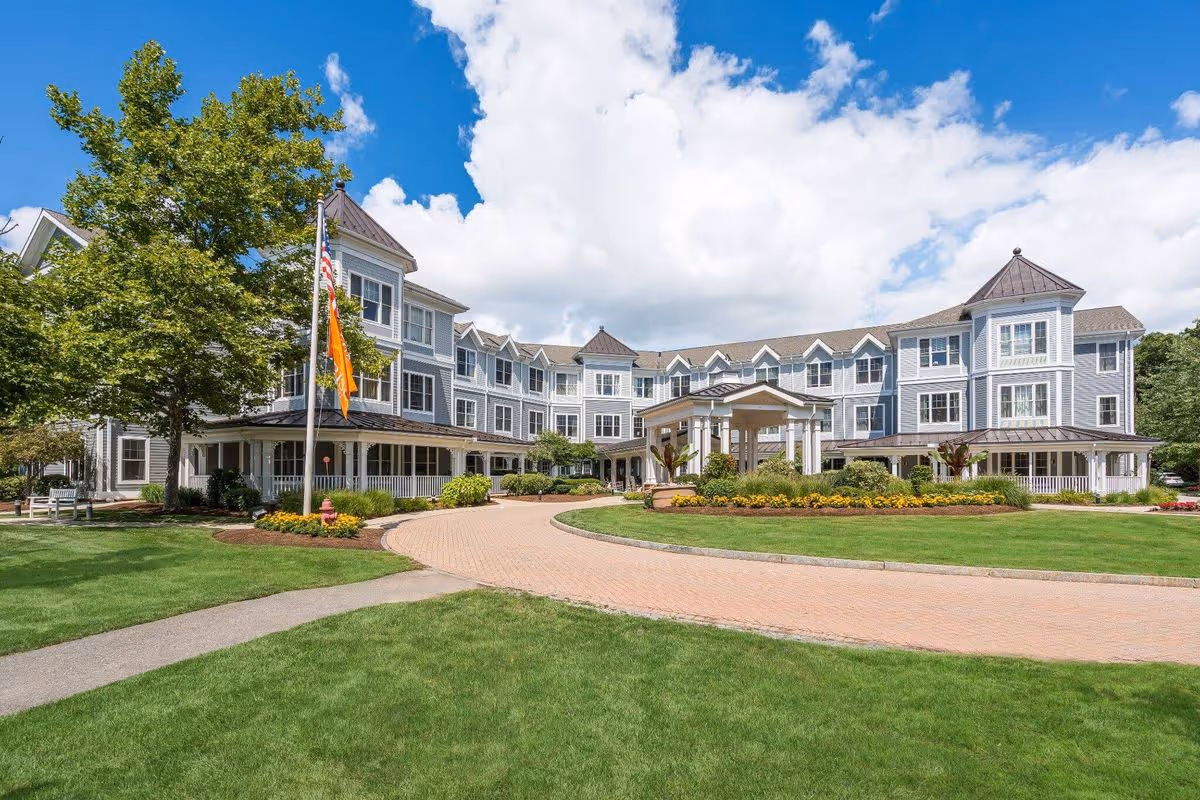 Exterior view of a large, three-story senior living facility building with gray siding and white trim, surrounded by well-maintained green lawns, a paved circular driveway, and landscaping with flowers and shrubs under a partly cloudy blue sky.