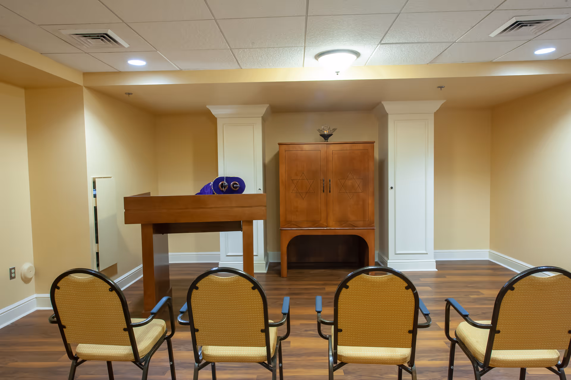 Small worship room with four chairs facing a wooden lectern and a cabinet marked with Stars of David.
