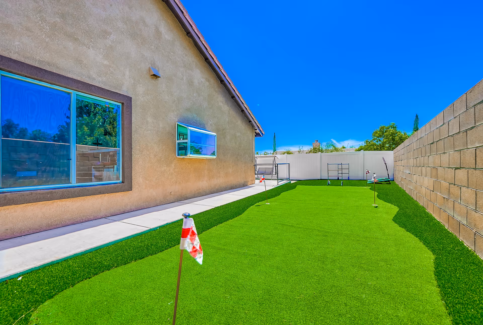 Outdoor backyard area with a small artificial putting green featuring several golf flags, adjacent to a beige stucco building with windows, surrounded by a concrete block wall and a white fence under a clear blue sky.