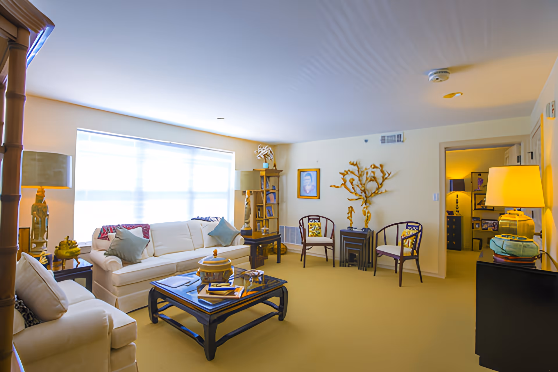 A bright and cozy living room with two white sofas, a dark wooden coffee table with books and decorative items, two wooden chairs with cushions, a tall lamp, and various decorative pieces including a golden tree sculpture and framed artwork on the walls. The room has a large window with blinds letting in natural light.