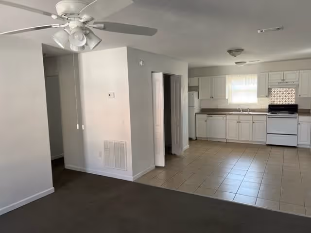 Open-plan interior showing a living area with a ceiling fan and an adjoining kitchen with white cabinets and tiled floor.