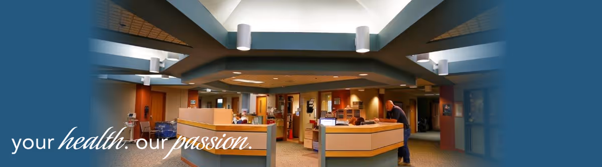 Interior view of a healthcare facility reception area with a central desk, staff working behind the desk, and a person leaning on the desk. The ceiling has modern lighting fixtures and the space is well-lit with a warm color scheme.
