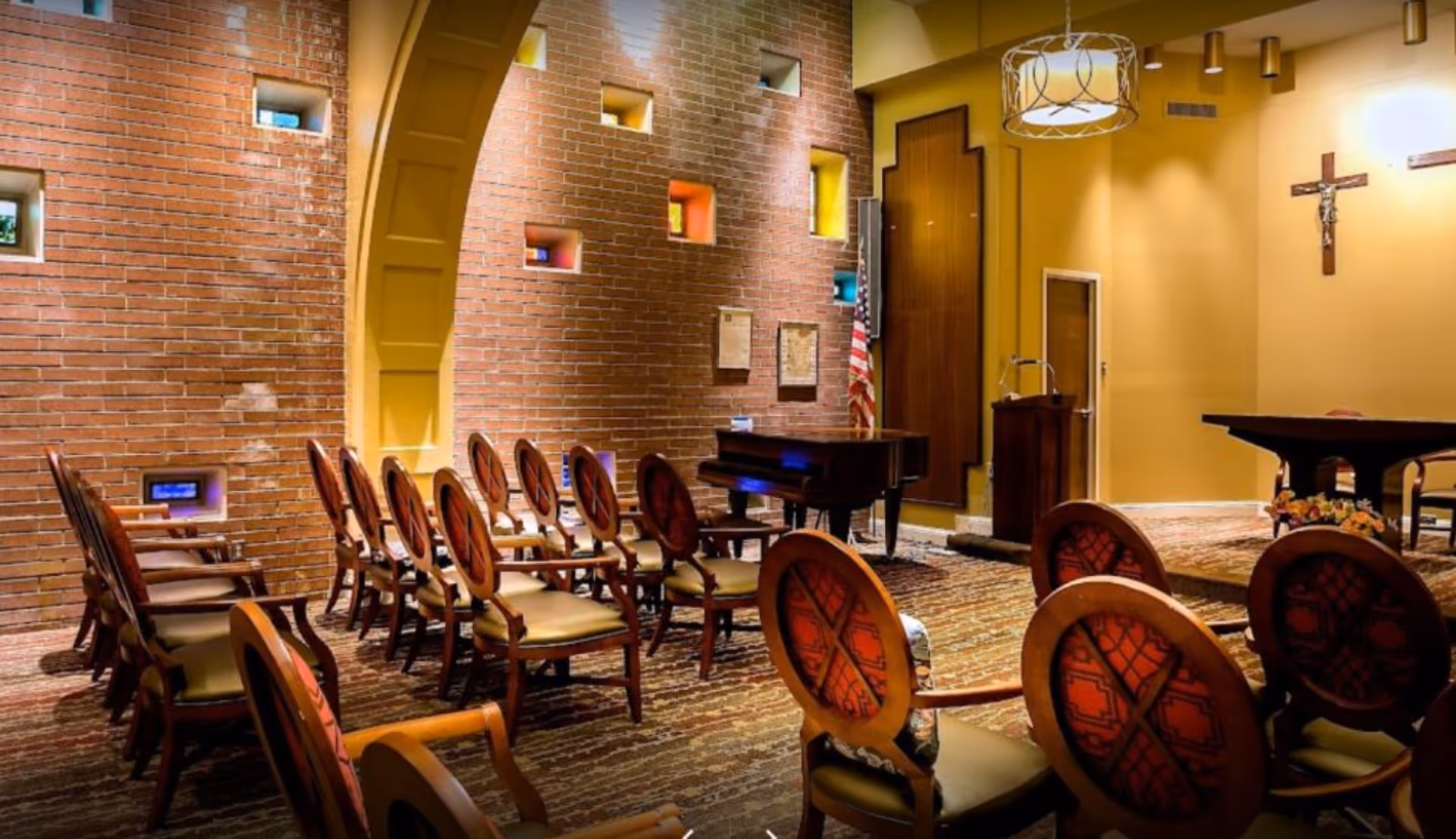 Interior chapel-style room with rows of wooden chairs, a piano, a podium, and a crucifix on the wall.