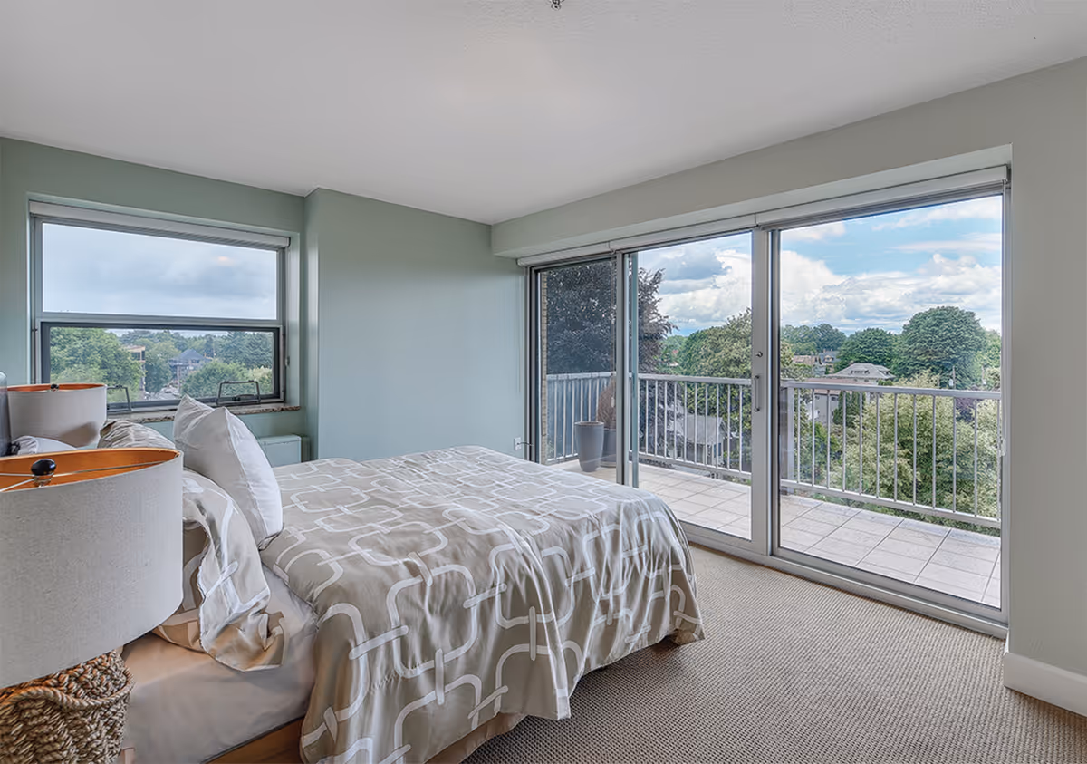 Bright bedroom with a made bed, nightstand lamp, and large sliding glass doors opening to a balcony with treetop views.