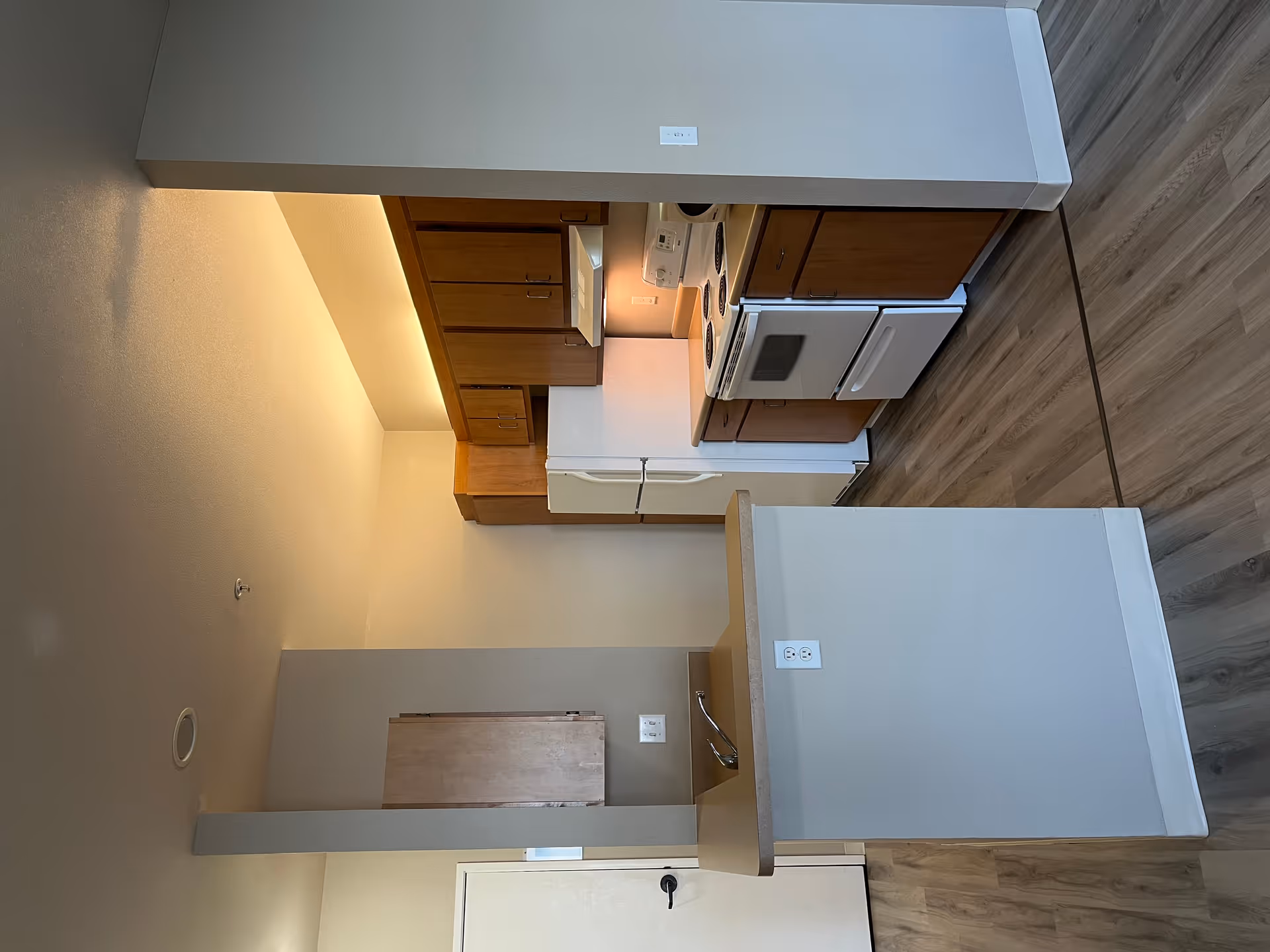 Interior view of a small kitchen area with wooden cabinets, a white refrigerator, a white stove with oven, and a countertop with a sink. The floor has wood-like vinyl flooring and the walls are painted light beige.