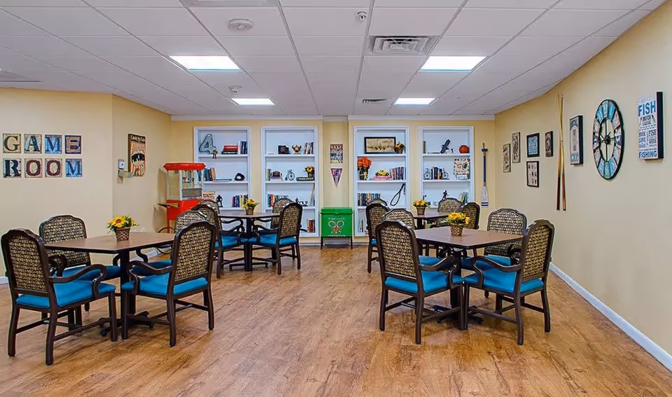 A game room with several tables and chairs arranged for group activities. The walls are decorated with various framed pictures, a large clock, and a sign that reads 'GAME ROOM'. There are built-in shelves filled with books and decorative items, and a popcorn machine is visible in the corner. The floor has a wood finish and the ceiling has recessed lighting.
